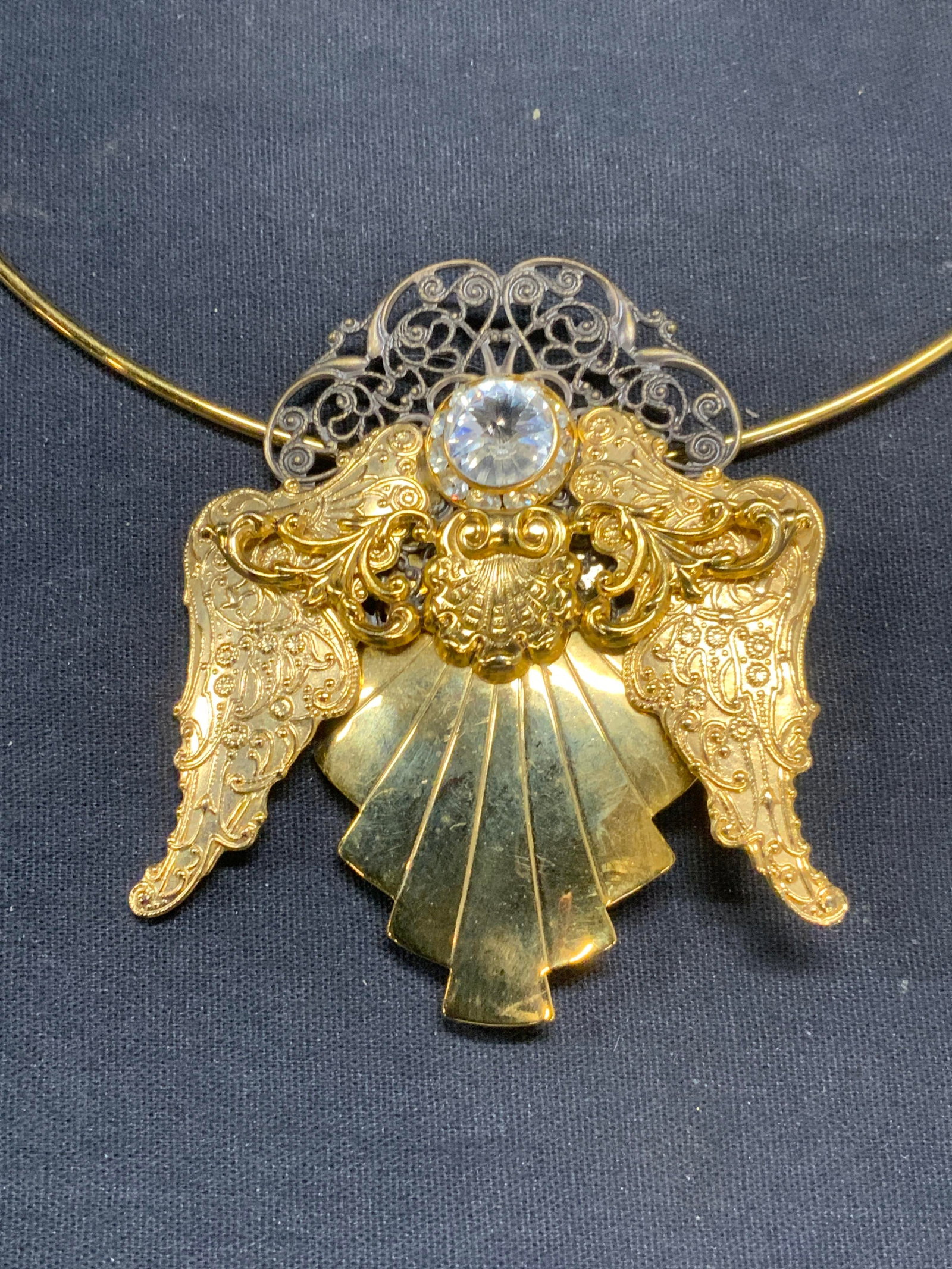 Gold Tone Brooch Pendant Necklace: Gold tone choker necklace with brooch pendant. Gold tone brooch with decorative ornate embossed detail and colorless crystal. Hinge pin to underside. Necklace measures approx. 5.25 inches wide. Brooch