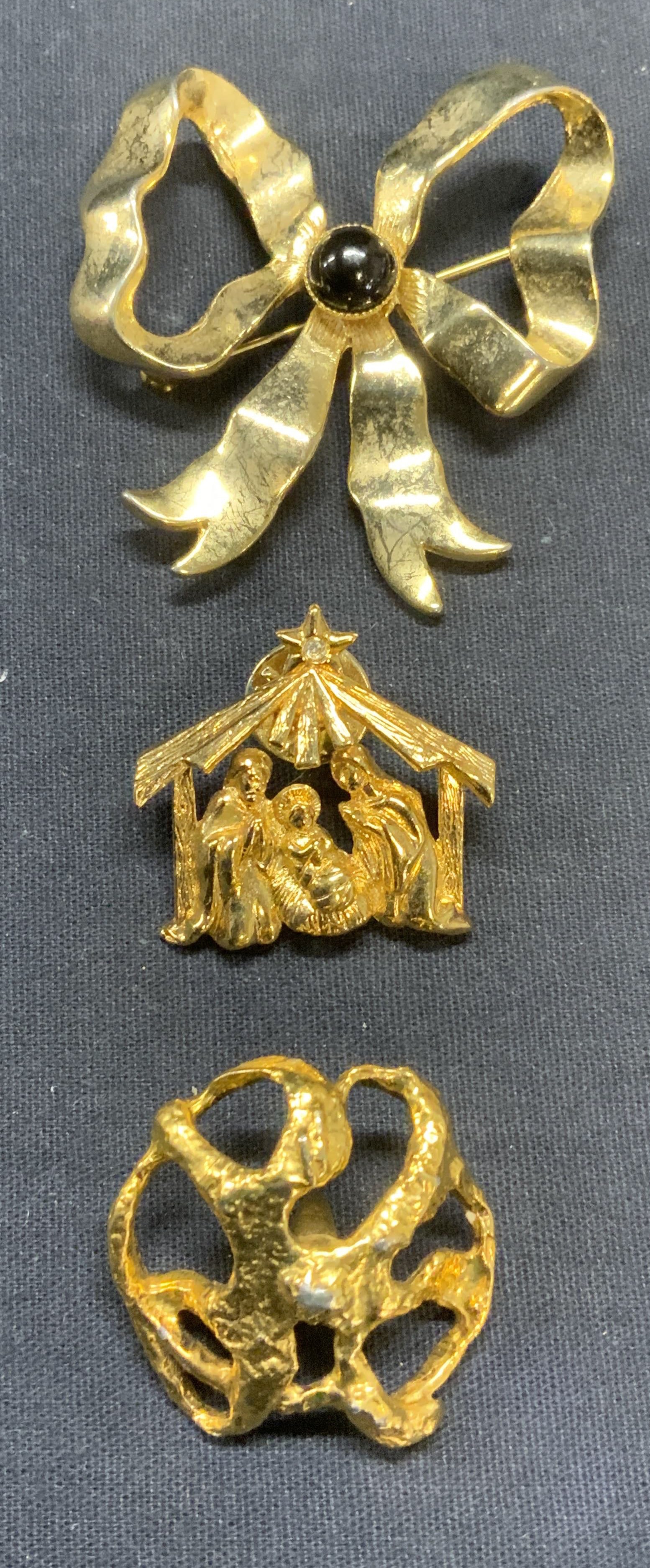 Lot 3 Gold Tn Ribbon Brooch, Holiday Pin +: Lot of 3 jewelry pieces. Gold tone and black cabochon ribbon bow brooch. Hinge pin to underside. Gold tone manger pin with Virgin Mary, Christ, and Joseph. Gold tone abstract sculptural pendant. Large