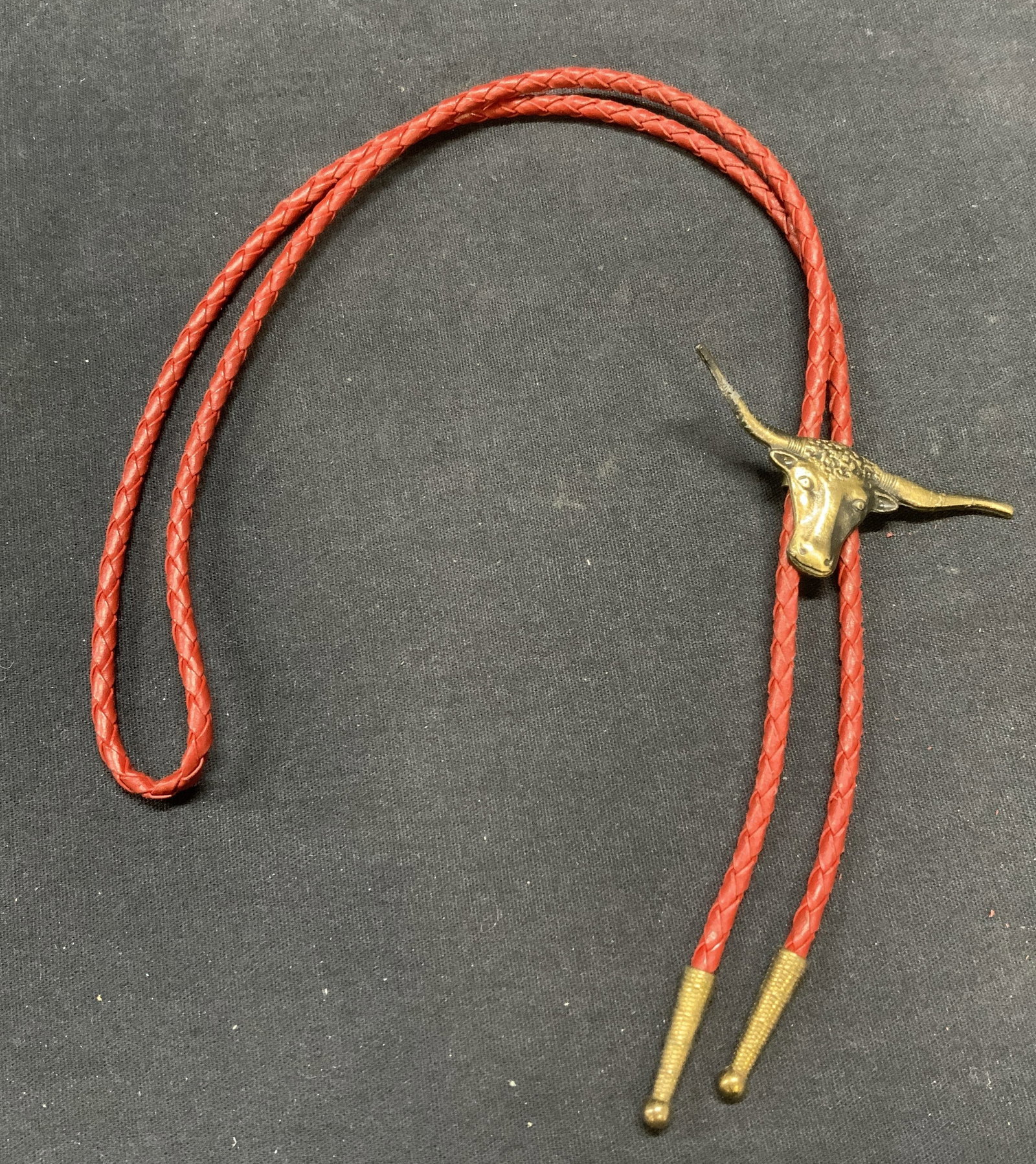 Gold Tone Red Leather Cow Bolo Tie Necklace: Woven red leather bolo tie with gold tone caps and longhorn cow motif. Drop length approx. 17 inches. Surface wear including scratching and bending to metal observed. Estate jewelry, costume jewelry, 
