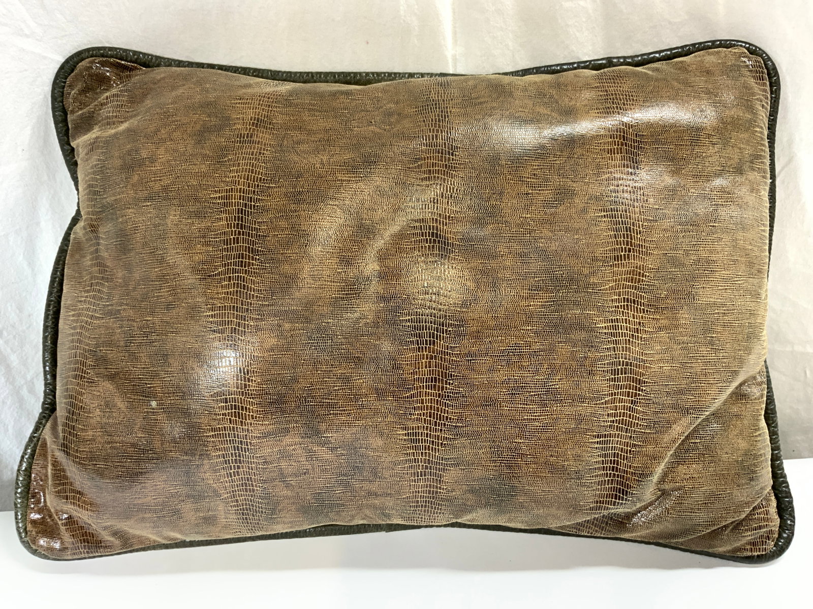 Croc Skin Style & Leatherette Welted Pillow: Pillow appears to be leatherette and croc skin style. Edges of pillow have thick welting, pillow is brown and slate green toned. Pillow measures approximately 24 inches wide 17.5 inches tall 6 inches 