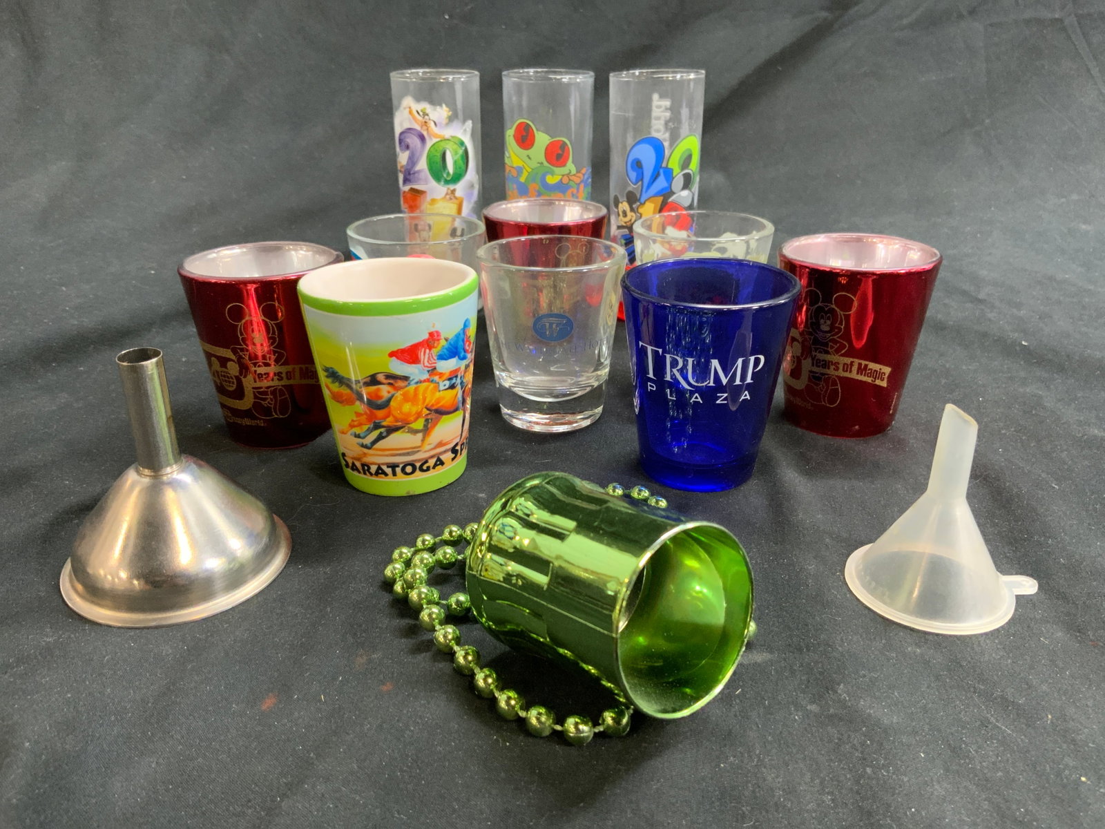 Lot 14 Barware, Disney, The Watergate Hotel & More: Lot includes 8 shot glasses, 3 double shot glasses, 2 funnels, & 1 green toned beaded necklace with beer mug pendant. Some trademarks include Disney, Trump Plaza, The Watergate Hotel, Saratoga Springs