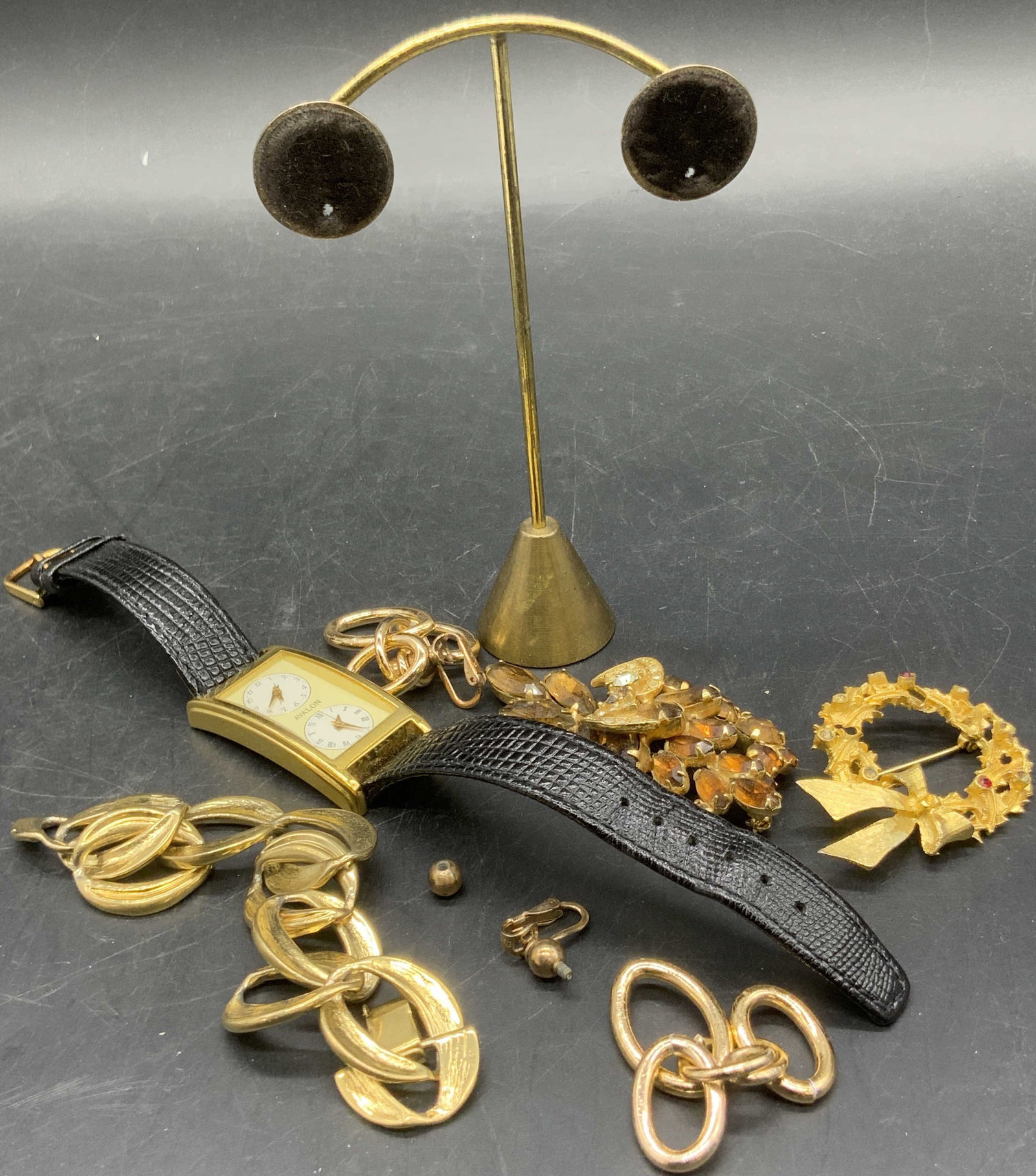 Jewelry Makers Lot, TRIFARI, AVALON & More: A jewelry makers lot consisting primarily of gold tone metal jewelry pieces and a metal earring stand. One watch featuring polished gold tone metal hardware and black leather band, face reads AVALON. 