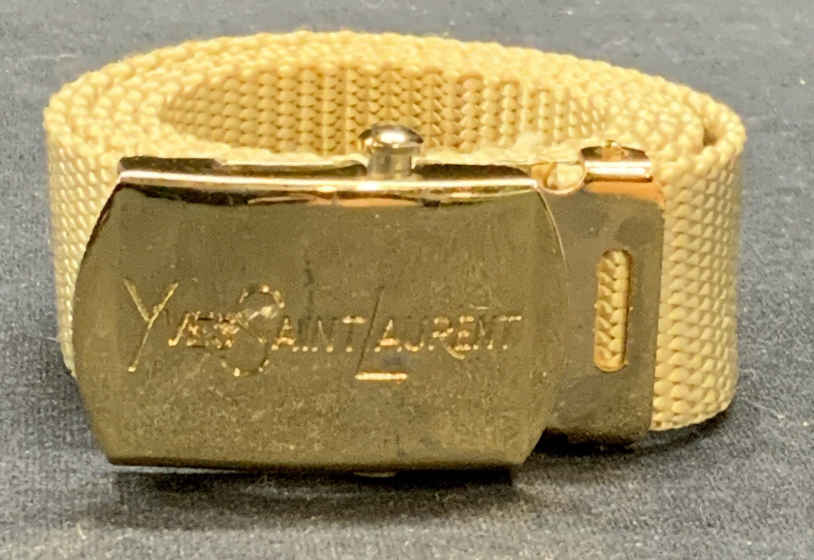 Yves Saint Laurent Logo Beige Belt: Yves Saint Laurent beige belt. Designer label on buckle. Textile and gold tone metal hardware. Measures approx. 35.5 inches long. Some surface wear consistent with exposure. YVES Saint Laurent, YVES S