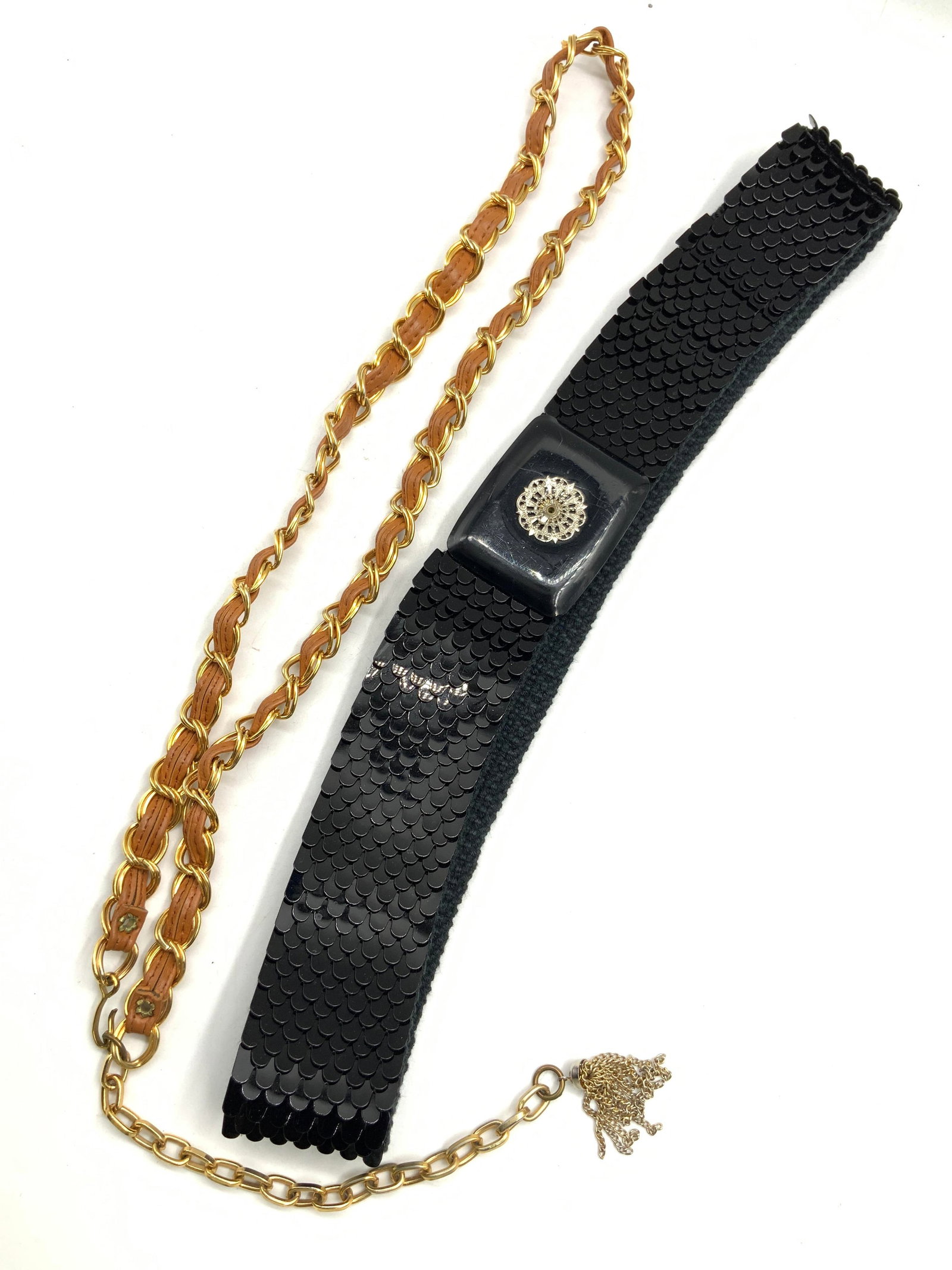 Lot 2 Leather & Woven Belts: Lot of 2 belts. 1 with black scale like sections made of metal with woven backing. The other a gold toned chain with brown leather braided through the chain and a tassel chain end. Sizes are 27 inches