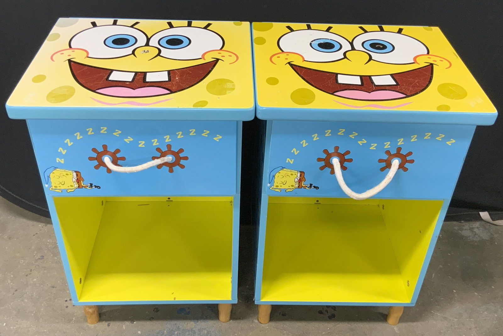 Pair SPONGEBOB SQUAREPANTS Nightstands: A pair of SPONGEBOB SQUAREPANTS motif nightstands. Crafted from wood and veneer with rope drawer pulls to the obverse. Tops feature SPONGEBOB SQUAREPANTS smiling while the obverse of the drawers pictu