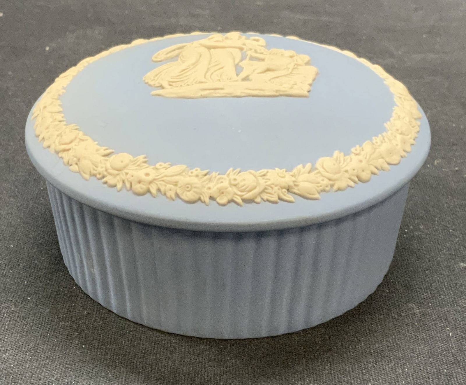 Vintage Wedgwood Blue Jasperware Box: Vintage Wedgwood blue Jasperware box. Round shaped with ribbed body. White bas relief Greco Roman motif with floral trim on lid. Trademarked to underside. Made in England. Measures approx. 3.5 x 1.25