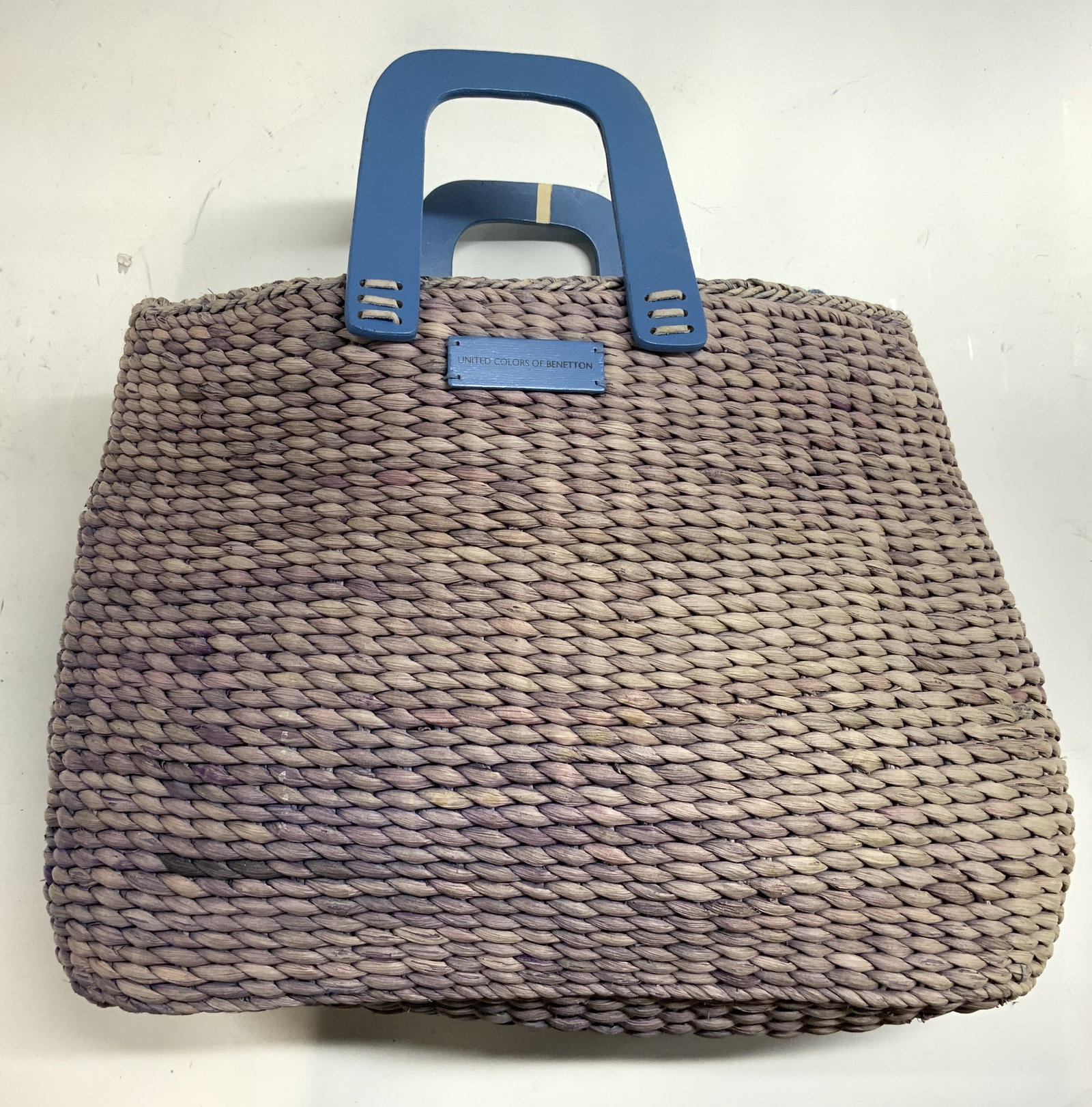 United Colors Of Benetton Woven Rattan Tote: Name plate on front reads UNITED COLORS OF BENETTON. Tote is woven rattan with light purple tone, blue toned interior and handles, zippered pocket inside. Handles appear to be wooden. Tote measures ap