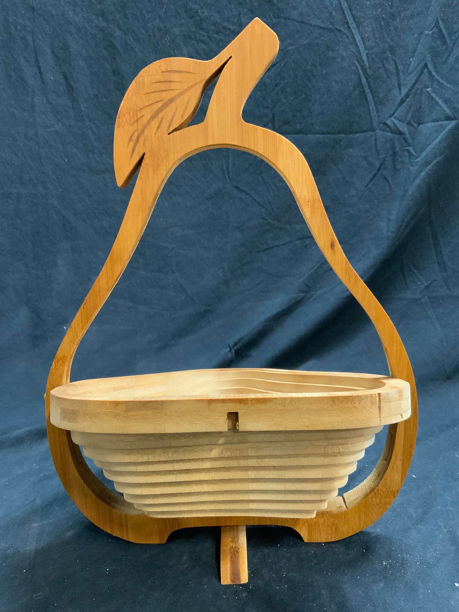 Collapsible Carved Wooden Pear Basket: Basket has pear form. Piece is tan toned and wooden. Basket rotates, collapses and expands. Base twists so basket can stand freely. Outer area has pear shape with leaf and stem form at top. Basket whe
