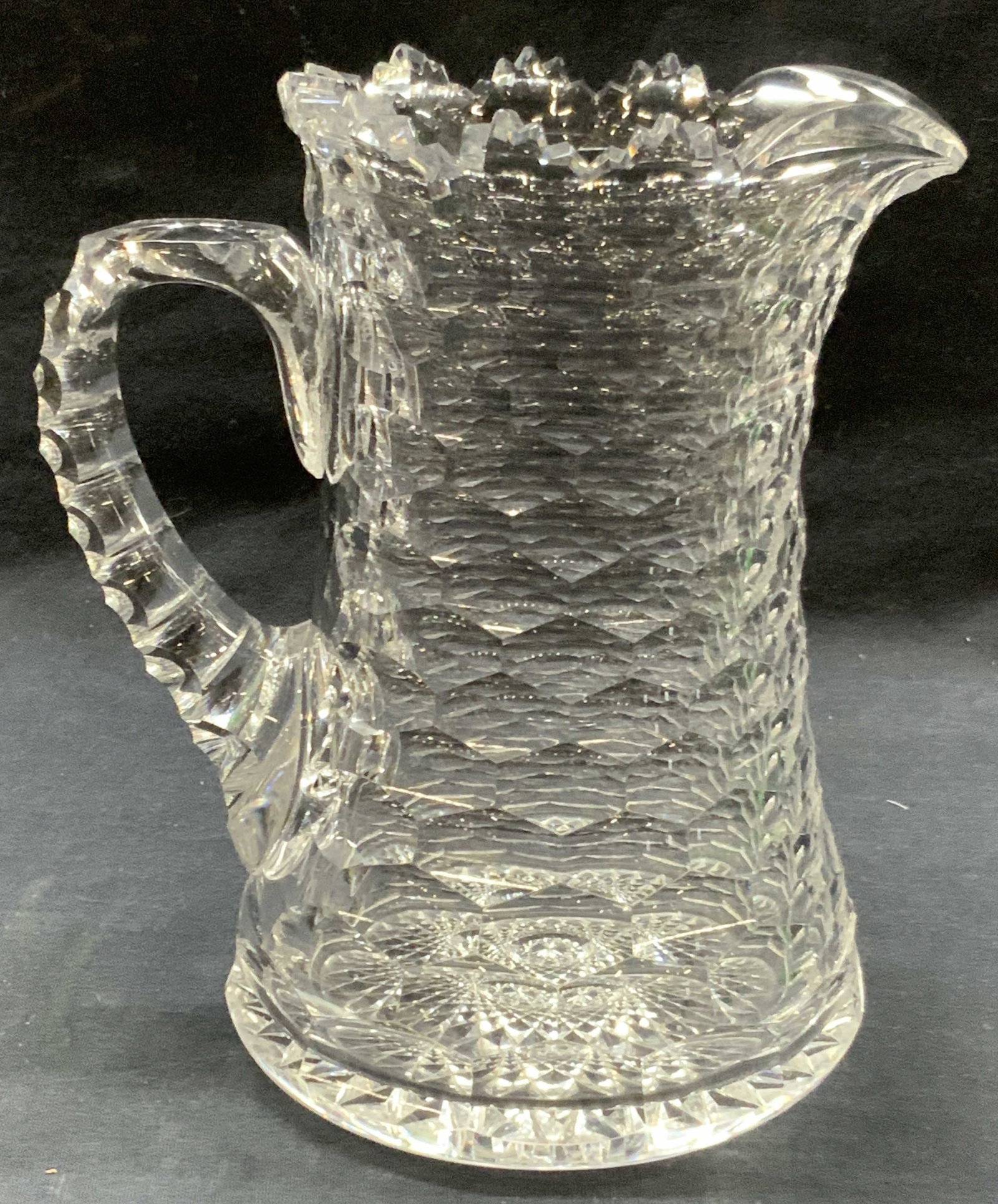 Hand Cut Clear Crystal Pitcher: Hand cut crystal pitcher with hexagonal shaped exterior patterns and saw tooth edges of pitcher with a lipped rim and back handles. Approx 8 inches tall and 4 inch diameter. Moderate wearing to crysta