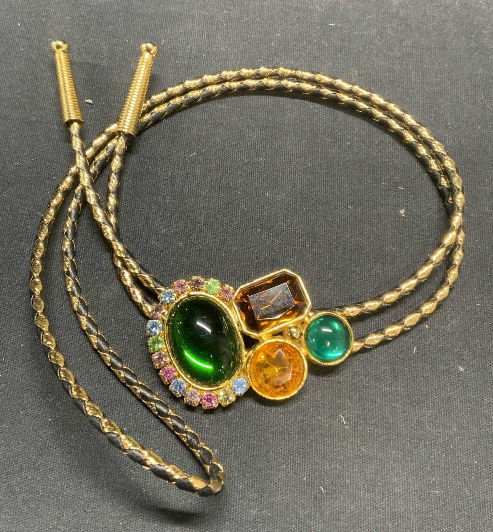 Gold Tone Crystal Bolo Tie Necklace: Gold tone and black braided cord bolo tie necklace with multi color crystal pendant. Drop length approx. 17.5 inches. Pendant measures approx. 2.25 x 1.75 inches. Some scratching observed. Estate jewe