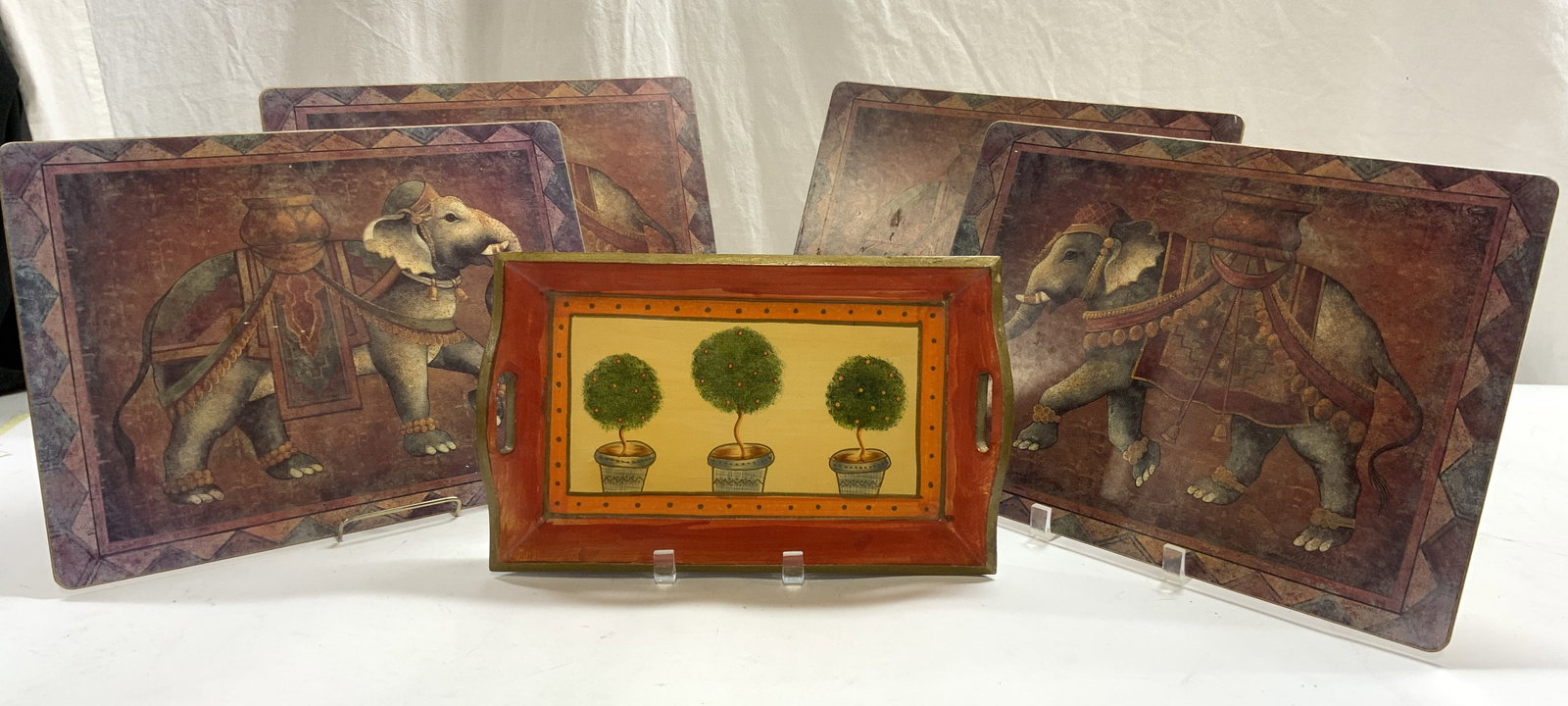 Lot 5 Set Of Elephant Placemats & Wood Tray: Lot includes a set of 4 placemats and 1 hand painted wooden tray. Each placemat is depict an elephant with vessel on back. Edges have geometric detail. Each placemat has rounded rectangle shape. Tray 