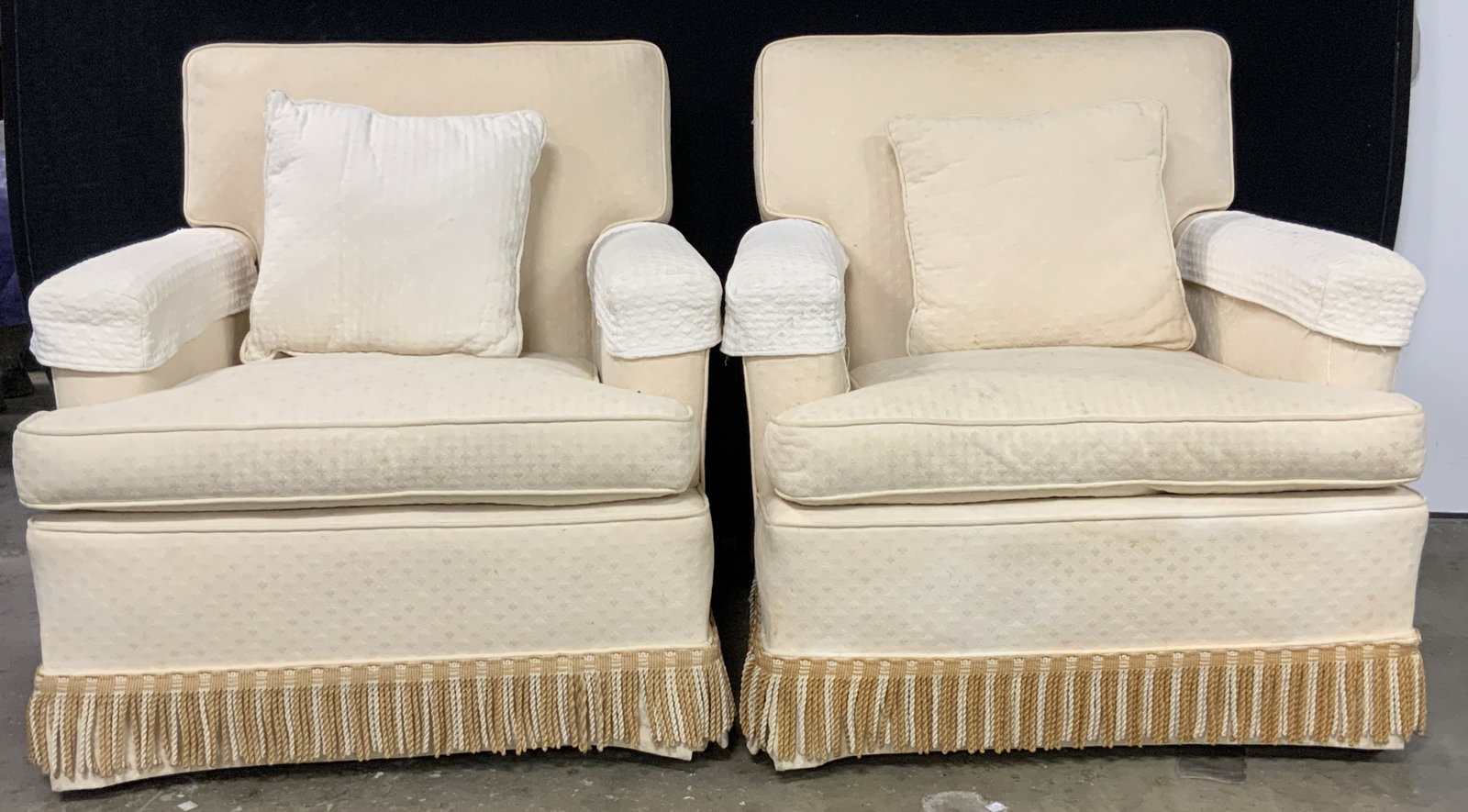 Pair Vintage Upholstered Armchairs W/ Toss Pillows: A pair of vintage upholstered armchairs. The chairs are upholstered in a cream hued upholstery. Tassel trim along the skirted bases. Chairs come with toss pillows which feature a solid colored side an