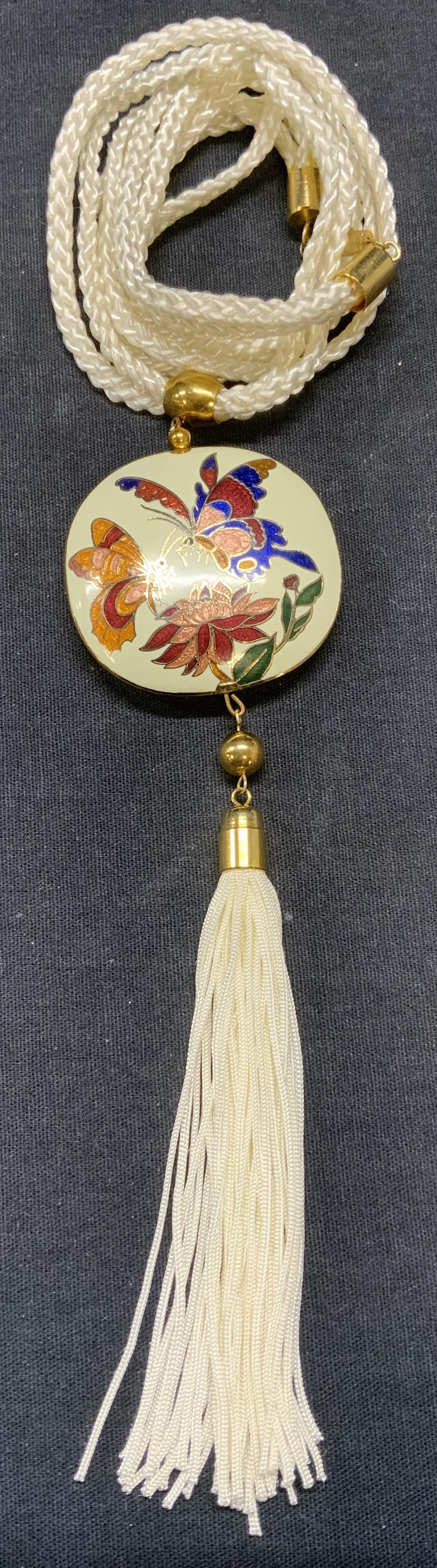 White Enameled Floral Pendant Necklace: White cord pendant necklace with enameled pendant with floral motif and tassel. Drop length approx. 13.5 inches. Pendant and tassel length approx. 6.75 inches. Some surface wear including scratching o