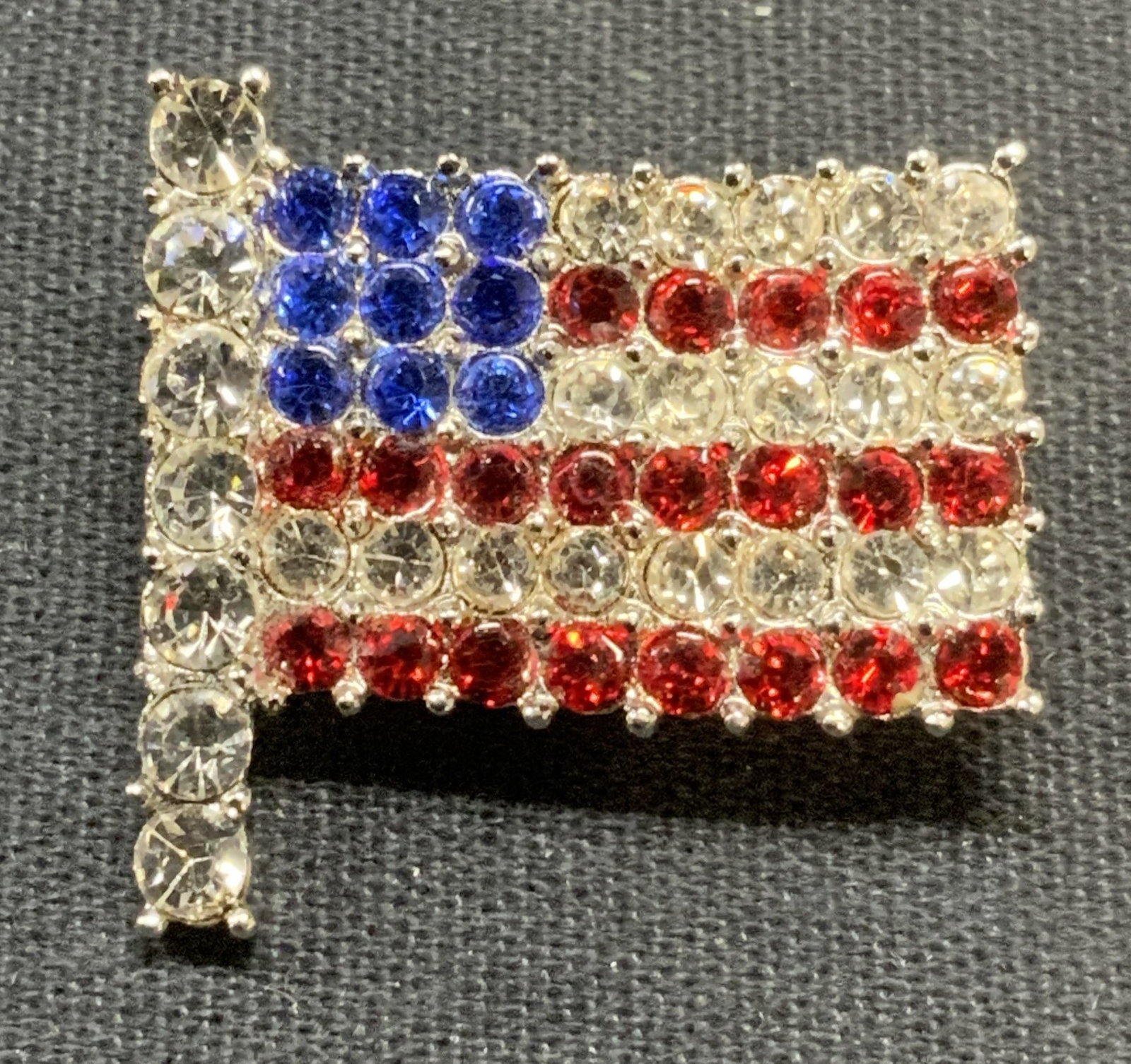 Silver Tone American Flag Crystal Brooch: Silver tone American flag brooch with colorless, red and blue crystal decoration. Hinge pin to underside. Measures approx. 1 x 1 inches. Some surface wear including scratching observed. Estate jewelry