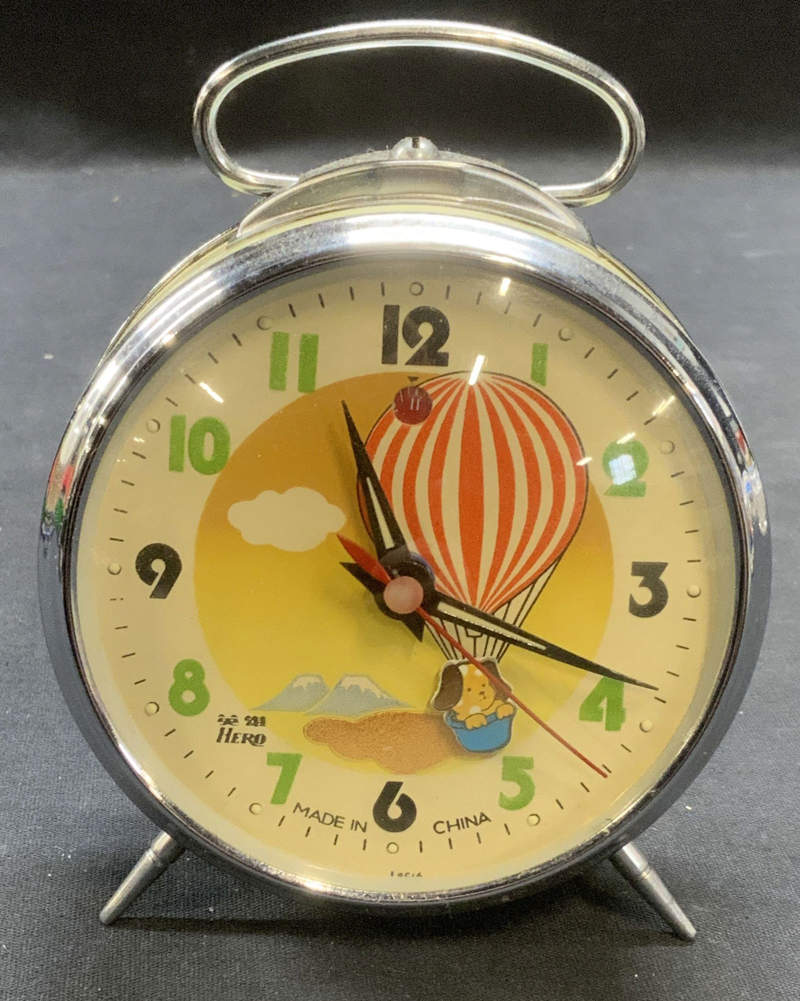 Vtg Hero Dog Hot Air Balloon Alarm Clock: Vintage Hero alarm clock with dog and hot air balloon motif. Trademarked on clock face. Composed of metal and plastic. Measures approx. 3.75 x 4.5 inches. Some surface wear including scratching observ
