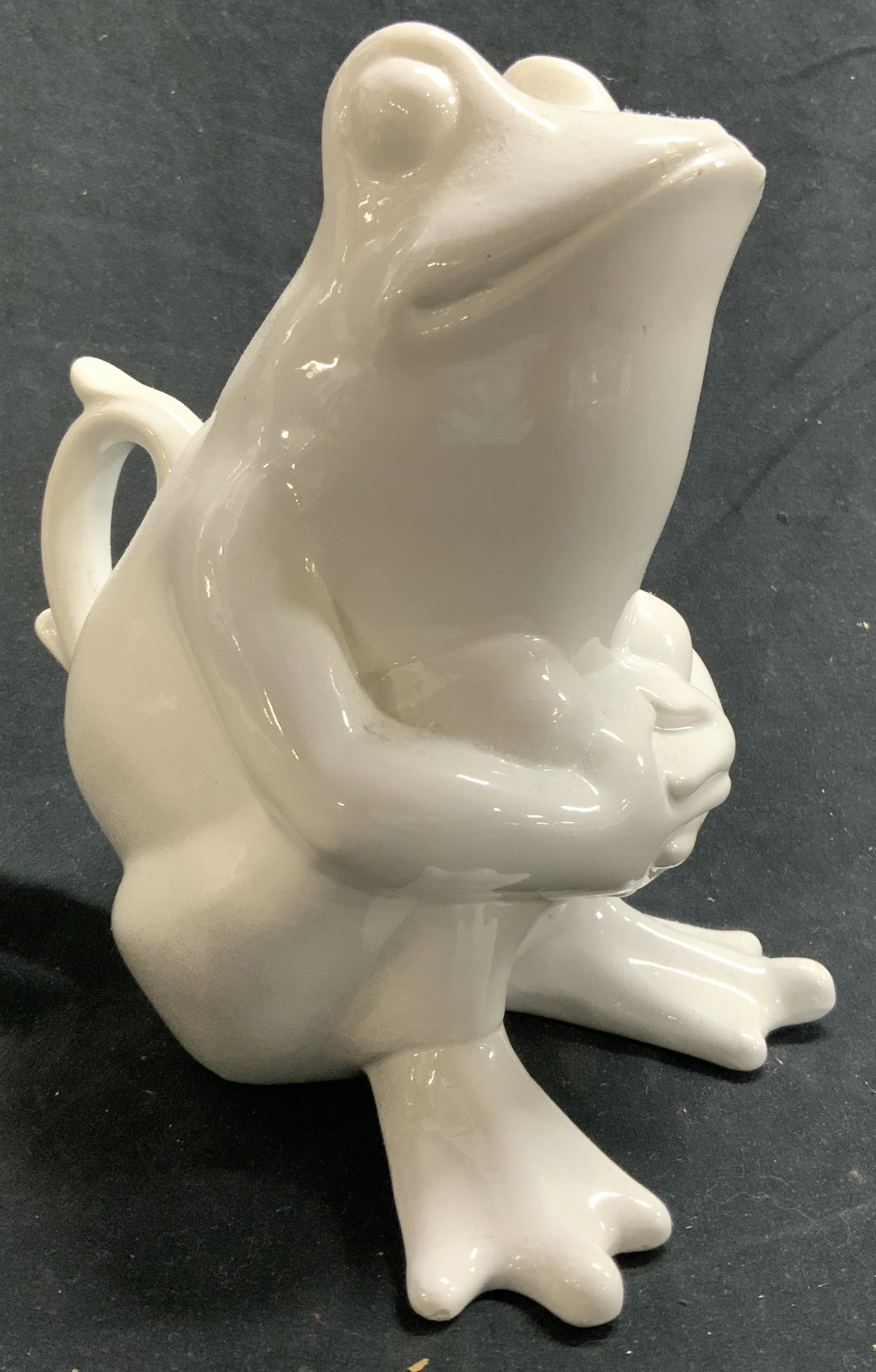 Porcelain Frog Pitcher Vessel White: Pitcher has seated frog form. Piece is white toned and porcelain. Pitcher handle has half heart form. Spout is at frog figures lips. Piece measure approximately 7 inches tall 6.5 inches deep 4.75 inch