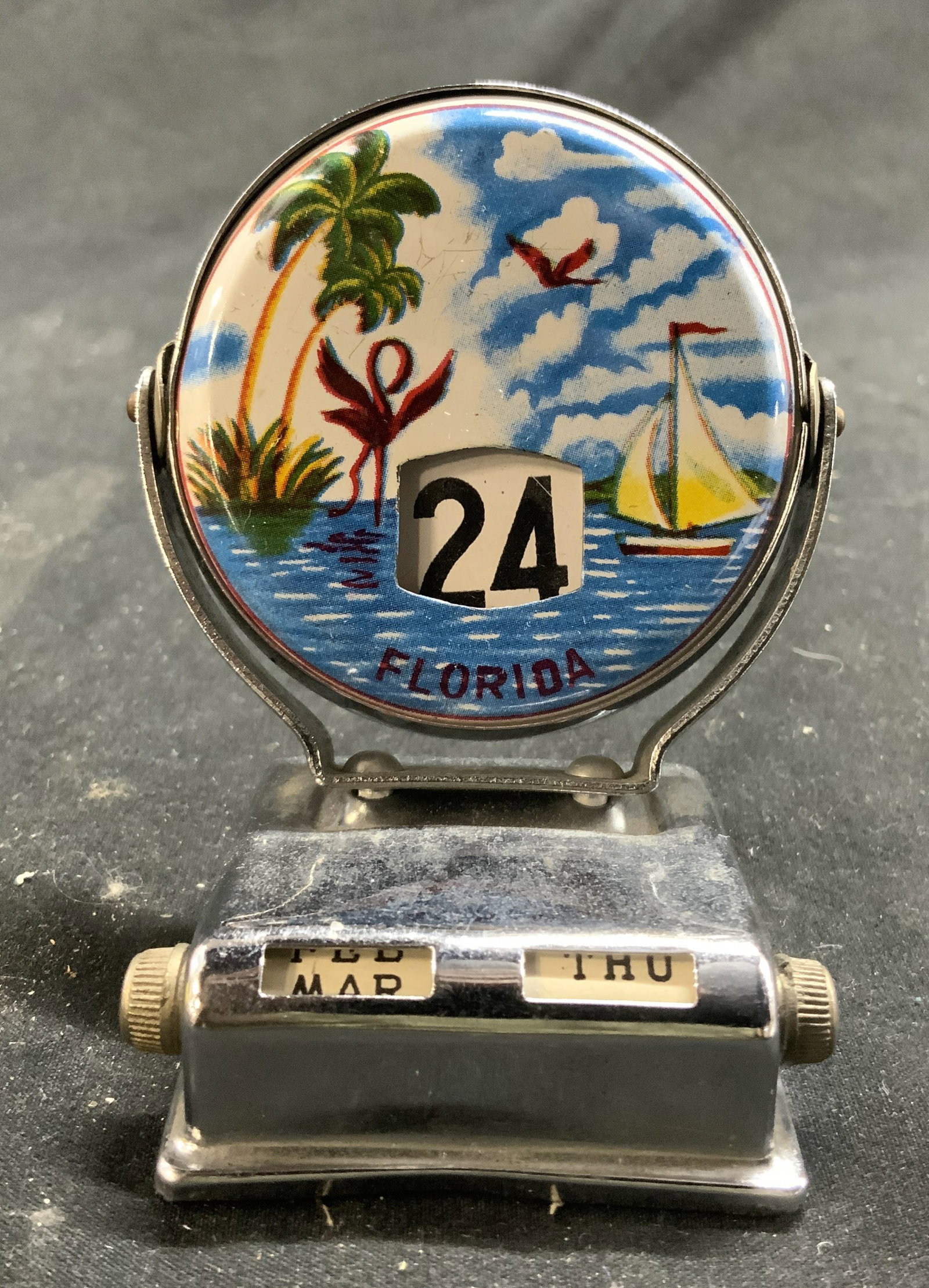 Vntg Florida Perpetual Flip Calendar: Calendar reads Florida. Piece has depiction of palm trees, boat at sea, birds and cloudy sky. Calendar has circular shape. Piece appears to be metal. Sides of base have twist knobs. Measures approx 3 