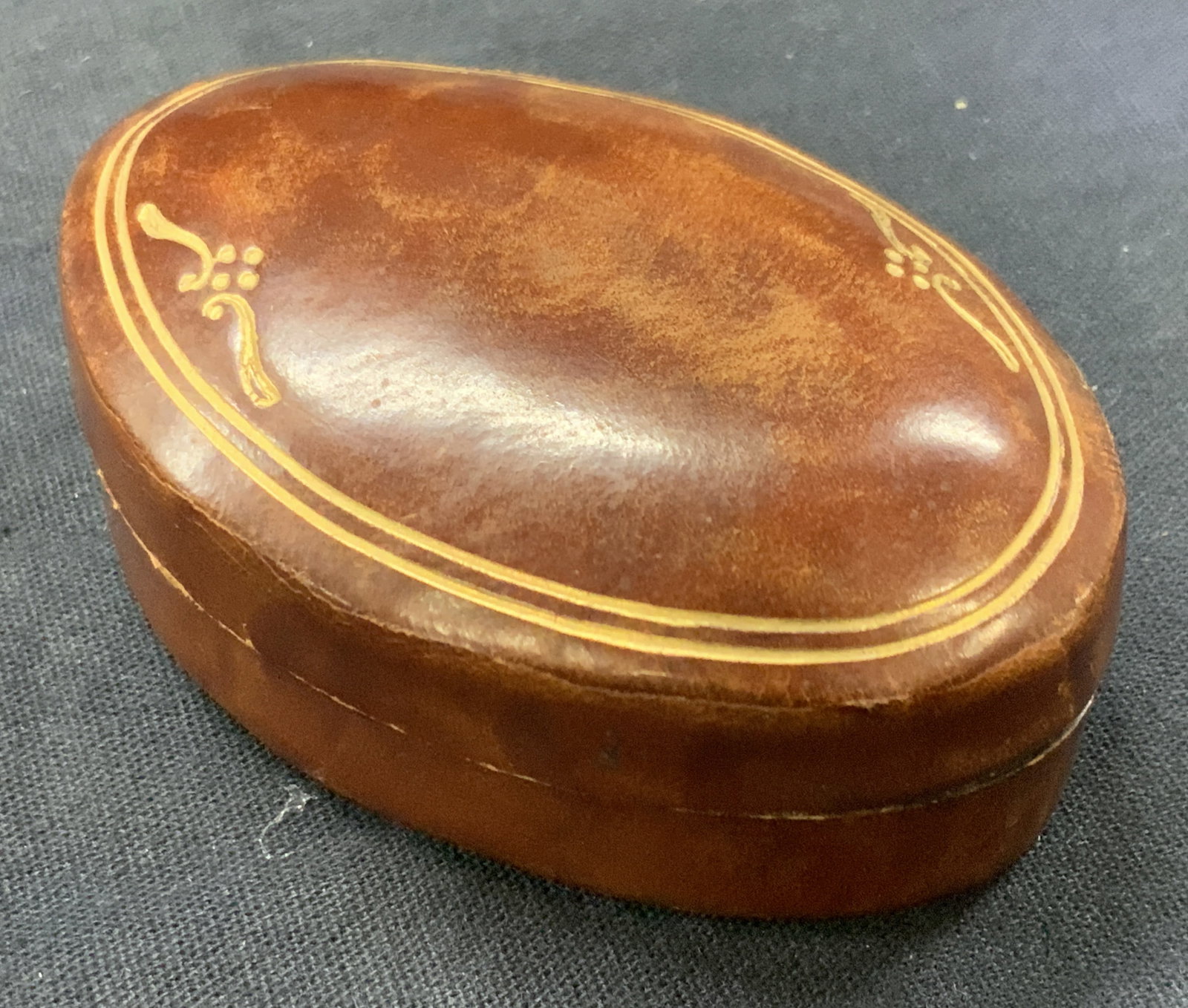 Gilt Leather Work Trinket Box, Italy: Leather made trinket box with gilt scroll designs on lid. Marked on underside Made in Italy. Brown toned in oval shape, approx 3 inches long and 2 inches wide. Moderate wearing to leather due to age a