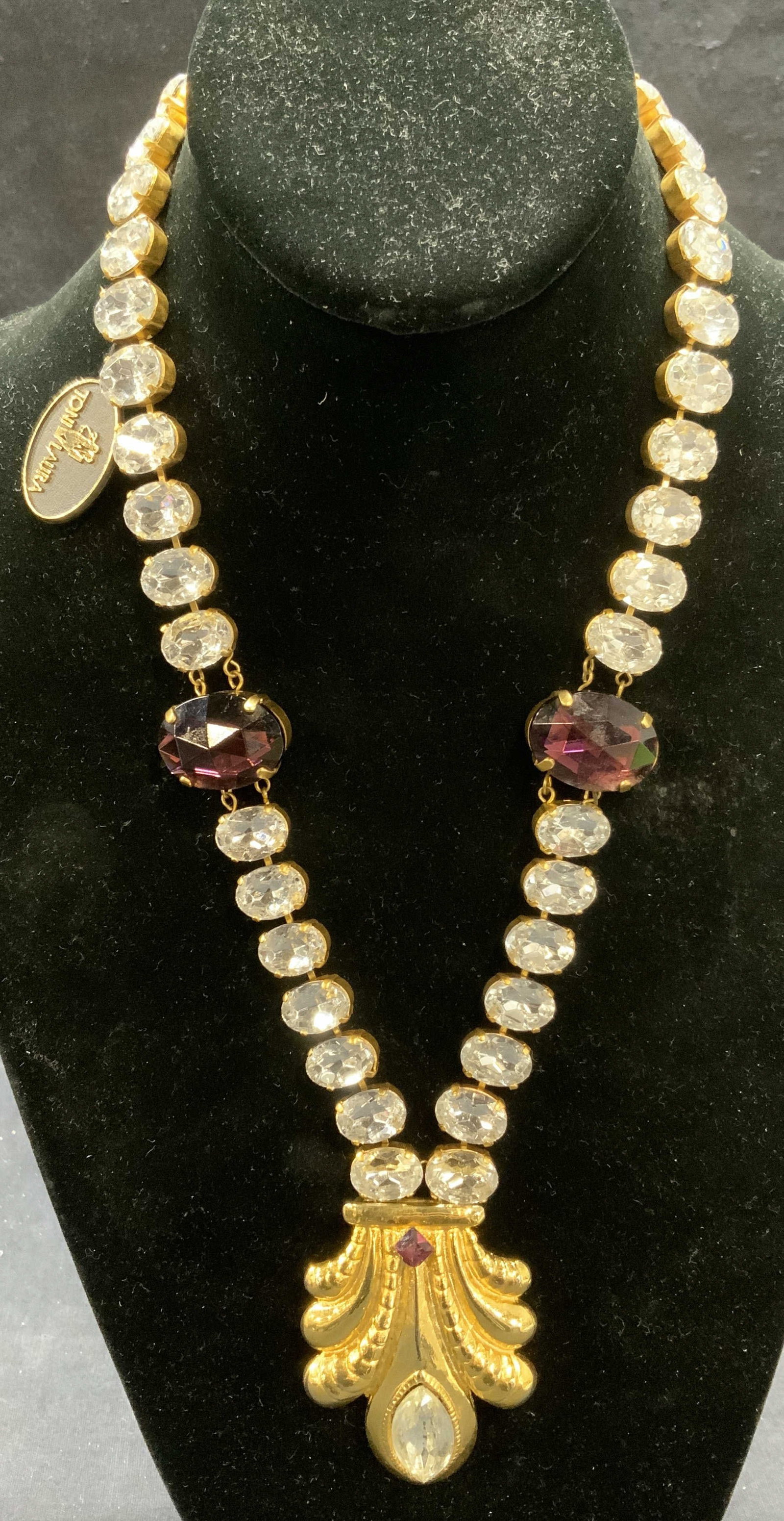 Vtg Toni Laura Gold Tn Crystal Pendant Necklace: Vintage Toni Laura gold tone pendant necklace colorless and purple crystal decoration. Embossed raised pattern on pendant. Signed on tag. Drop length approx. 10 inches. Pendant measures approx. 2.5 x 