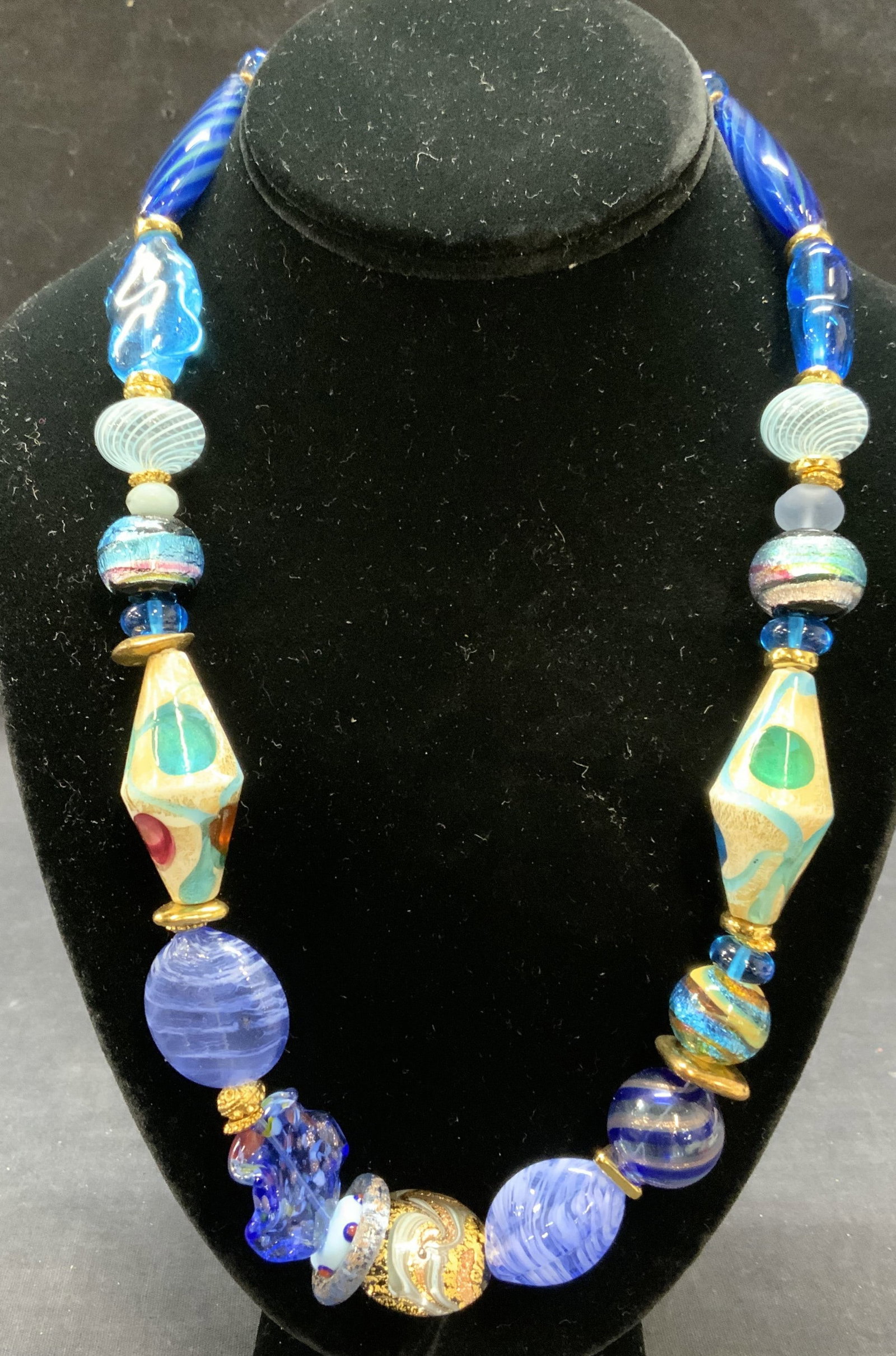 Gold Tone Blue Art Glass Beaded Necklace: Blue art glass beaded necklace with gold tone accent beads. Textured glass beads and swirl motif beads. Drop length approx. 10.5 inches. Some surface wear including scratching observed. Estate jewelry