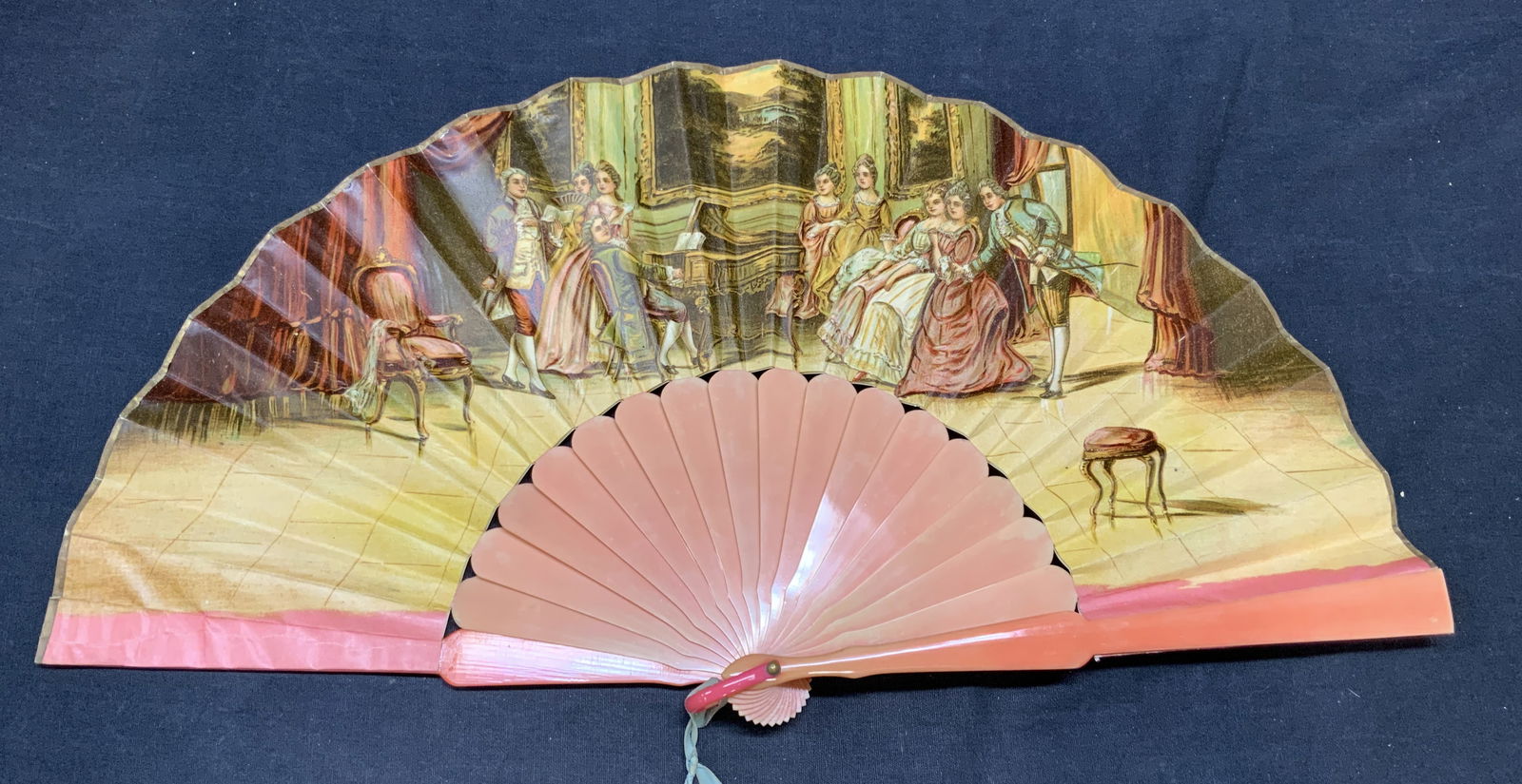 Antique Pink Figural Fan: Antique fan with Figural motif. Pink frame with printed interior scene with figures and piano. Paper and possibly cellulose. Measures approx. 10 inches long. Some wear to material consistent with expo