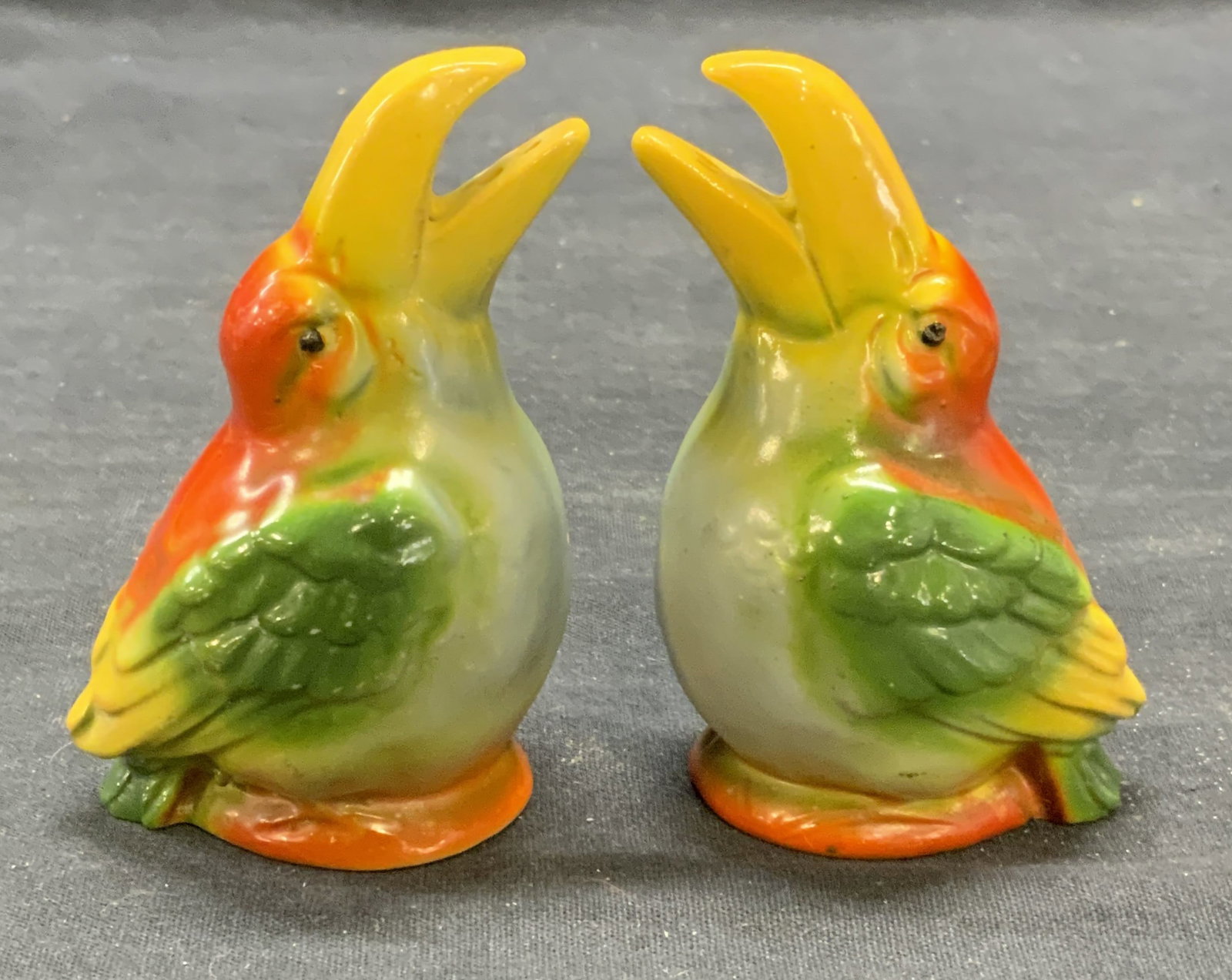 Pair Vintage Porcelain Toucan Shakers, Germany: Pair of vintage porcelain salt and pepper shakers. Toucan figural motif. Hand painted in hues of orange, yellow, and green. Measure approx. 2.25 x 3.5 inches each. Both pieces have some surface wear i