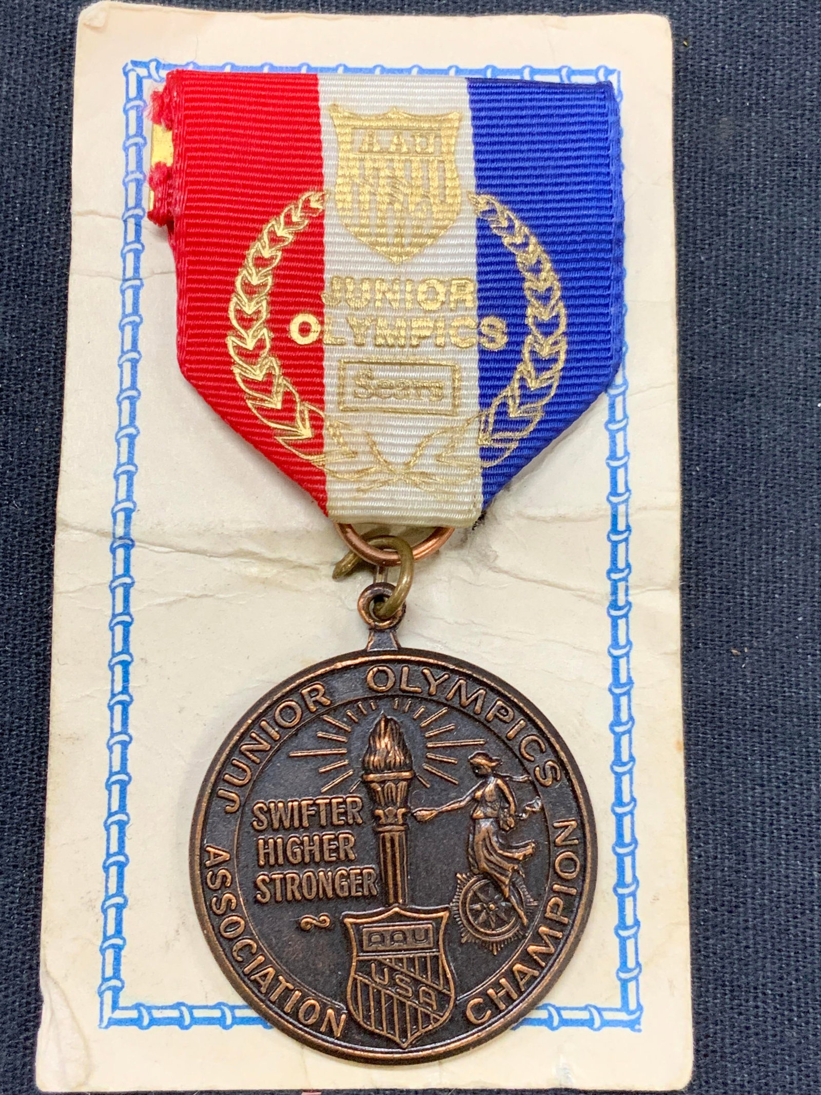 Copper Junior Olympics Badge, Sears.: Vintage copper badge with red white and blue ribbon, marked Junior Olympics, seats. Badge read Junior Olympics Association Champion. Approx 3.25 inches long and 1.25 inches wide. Vintage badge, copper