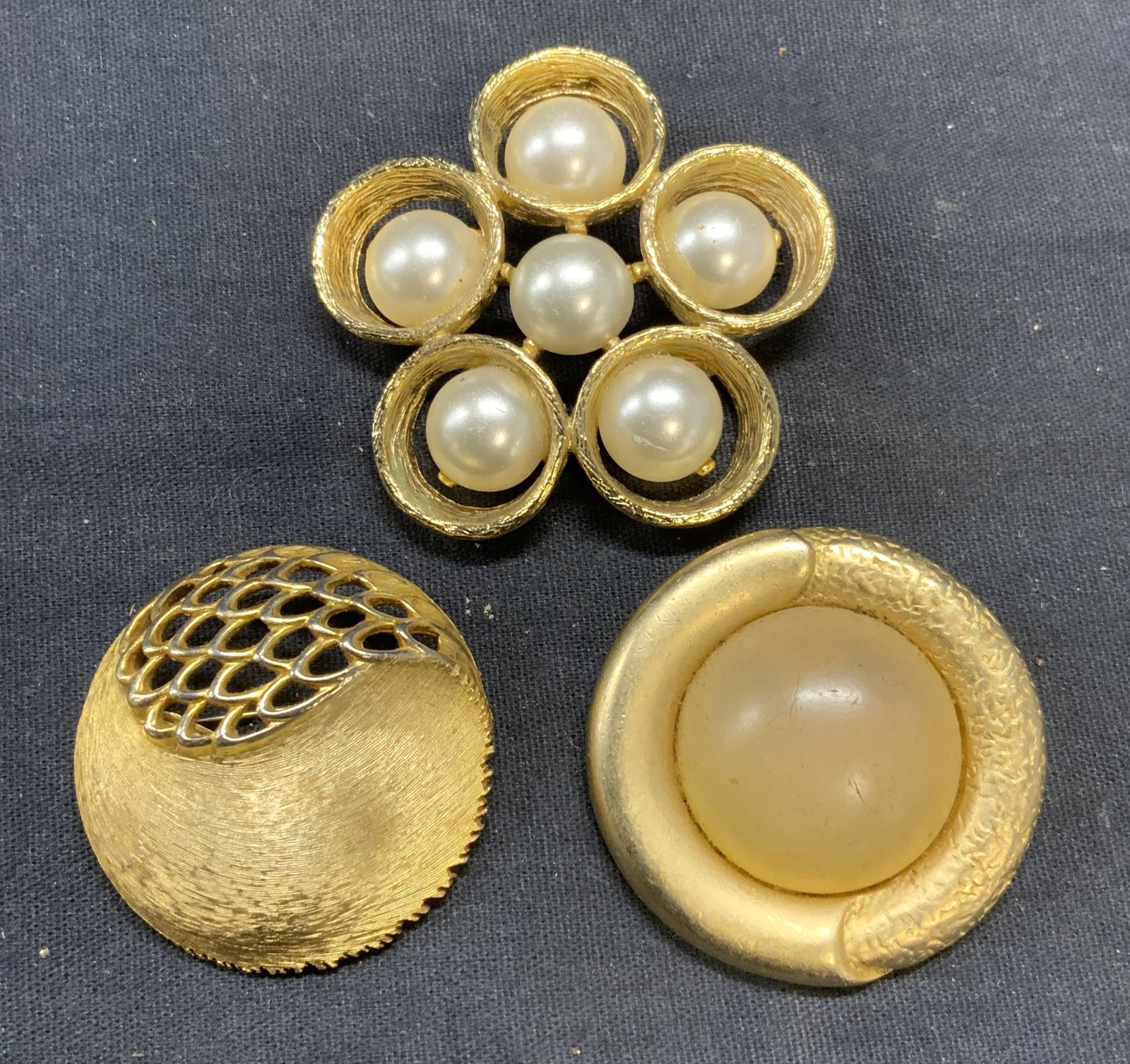 Lot 3 JJ & More Gold Tone Brooches: Lot of 3 gold tone brooches. Signed JJ brushed gold tone brooch with pierced detail. Signed with butterfly hallmark round cabochon brooch. Faux pearl flower brooch. All pieces have hinge pins to under