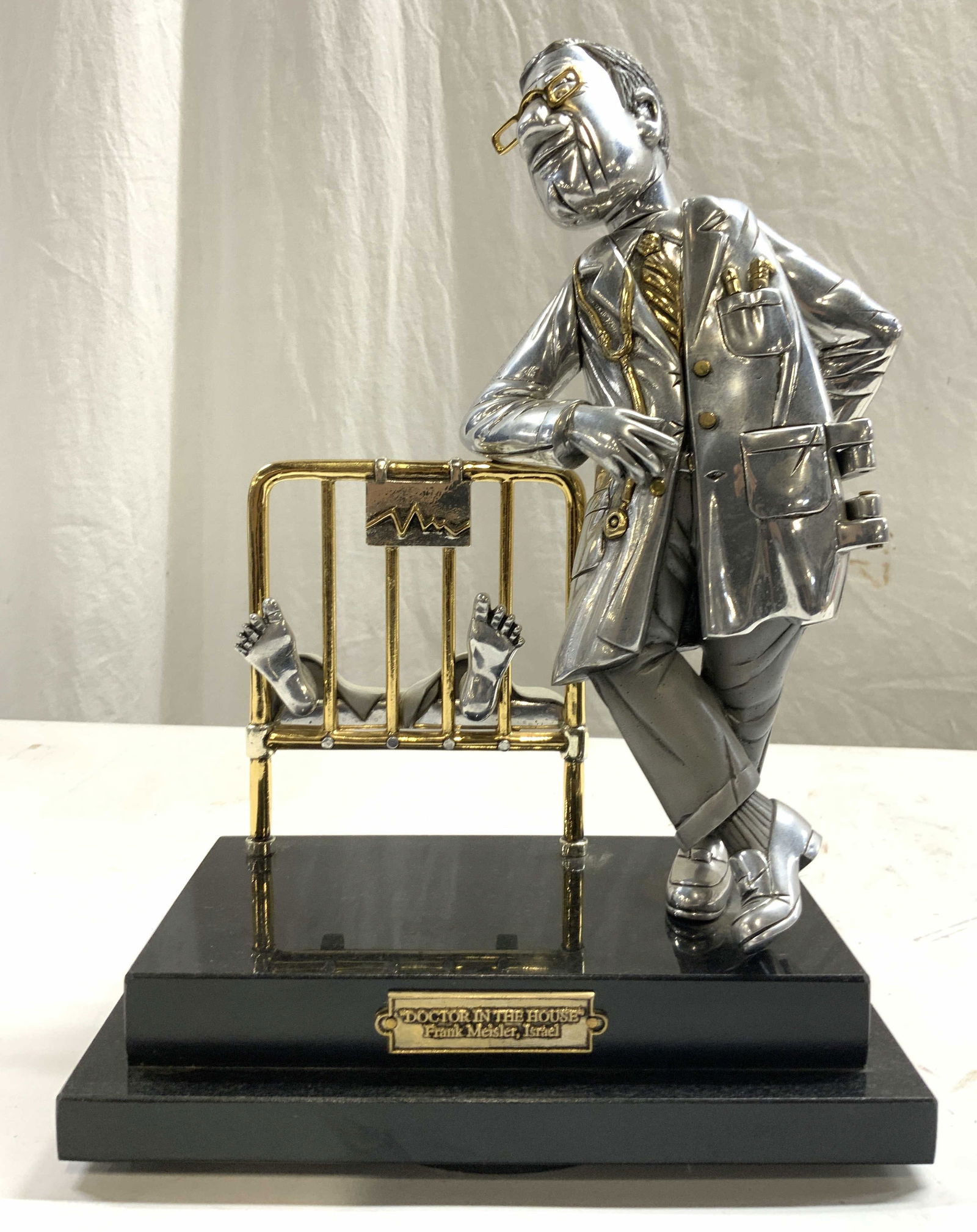 FRANK MEISLER Doctor In The House Metal Figural: Plaque on base reads FRANK MEISLER DOCTOR IN THE HOUSE ISRAEL. Sculpture appears to be pewter and gilt metal. Piece has black toned marble base. Depicts a male doctor figure with syringe behind back &