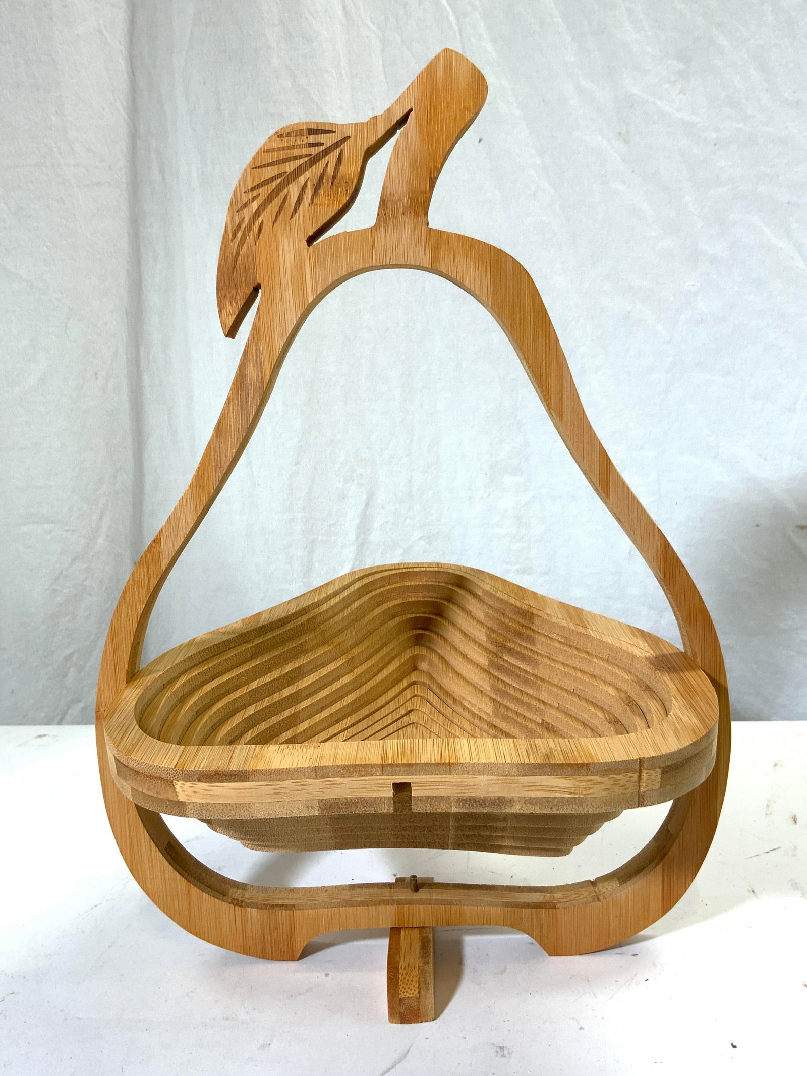 Collapsible Wooden Pear Form Basket: Basket is tan toned and wooden. Piece is pear shaped with stem and leaf at top with. Base of piece has twisting area to help basket stand on its own. Basket measures approximately 13.5 inches tall 10 