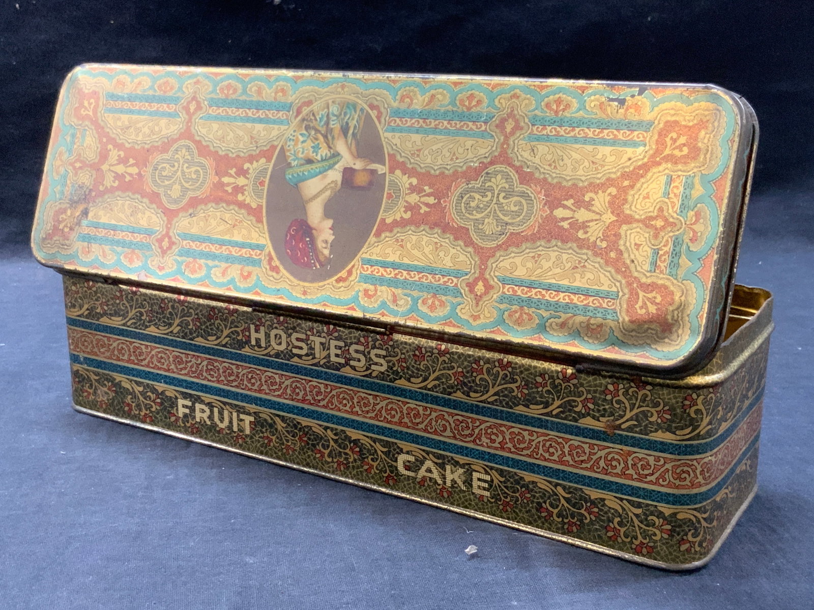 Vtg Hostess Fruit Cake Tin: Vintage Hostess fruit cake tin with art nouveau designs in tones of reds, blues and golds with a center image of a woman holding a fruit cake on a platter on the lid. Interior is adorn in gold tone. A