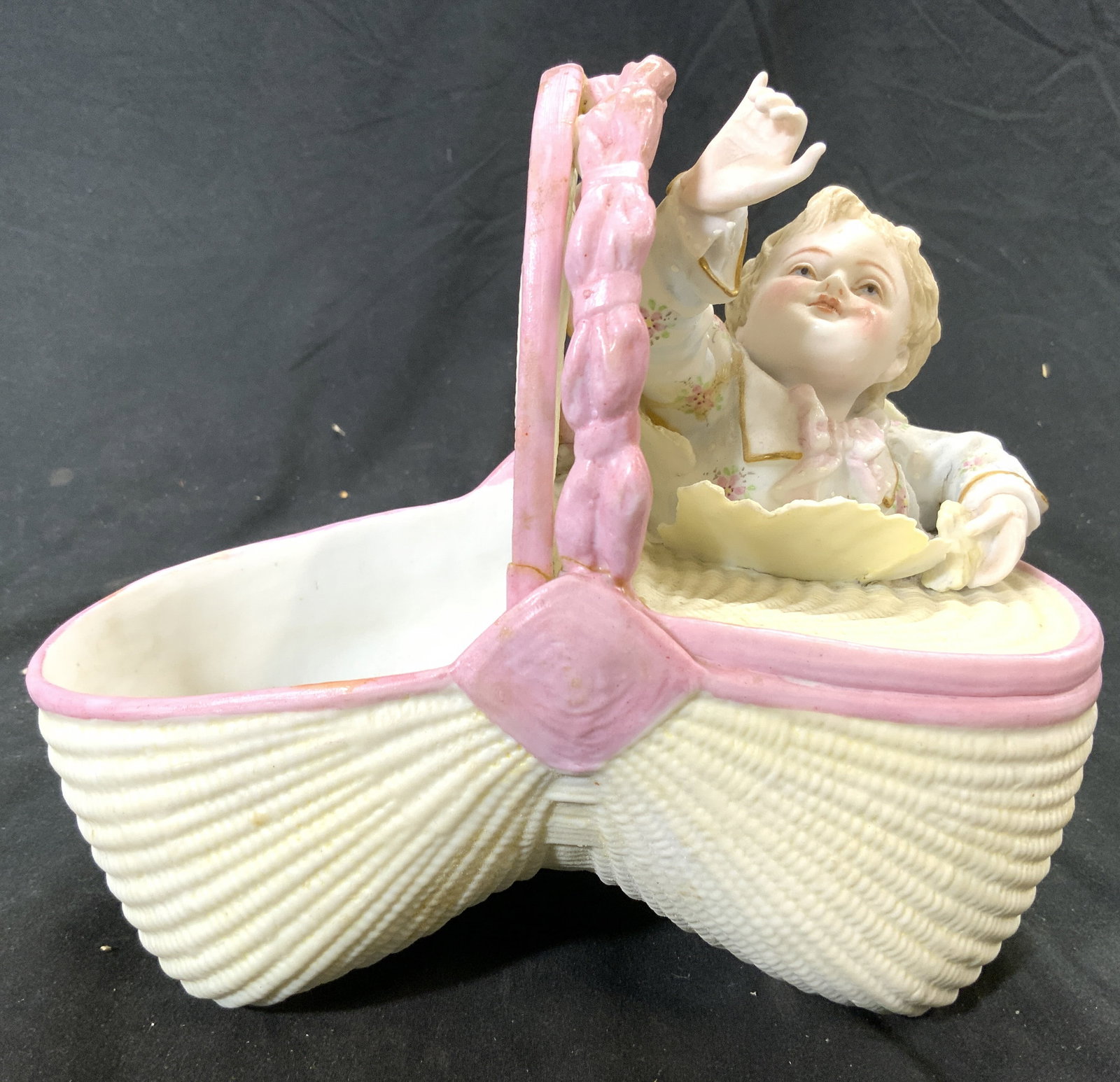 Vntg Porcelain Baby In Basket Figural: Figure depicts a baby in a woven basket. Piece is pink and yellow toned with white toned interior. Piece measures approximately 8 inches wide 7 inches tall 4.5 inches deep. Porcelain figure, porcelain