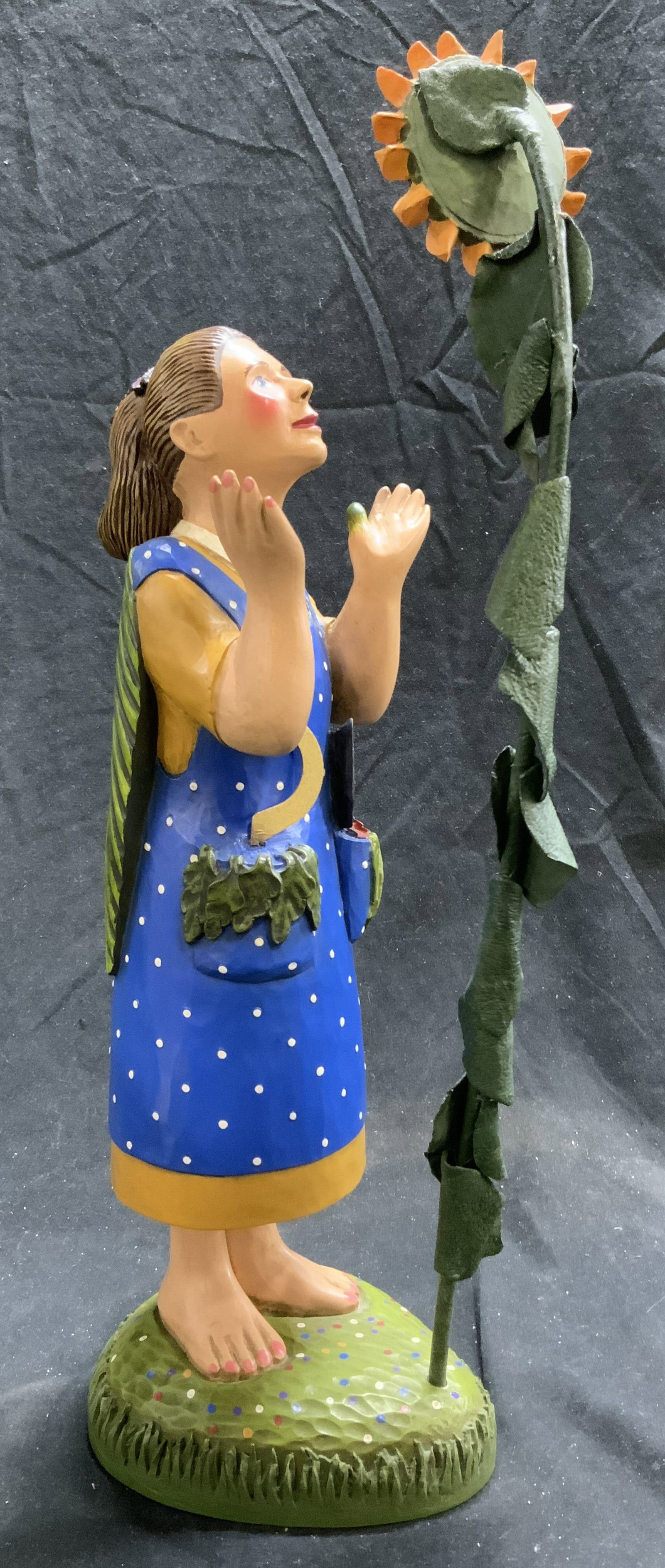 LEO R SMITH MIDWEST Carved Wooden Female Figural: Base is signed LEO R SMITH. Tag on underside reads MIDWEST. Edition 906/2500. Sculpture depicts a female figure wearing blue polka dot dress standing in front of large sunflower. Piece measures approx