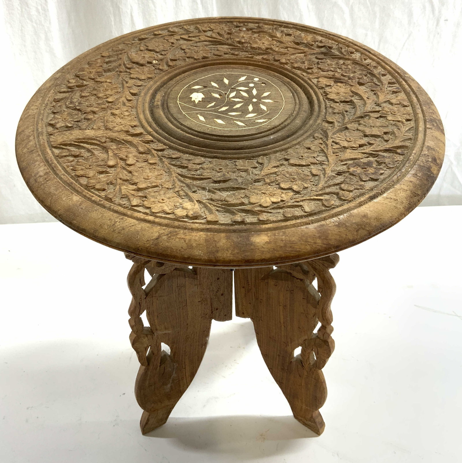 Vtg Anglo Indian Carved Wooden Folding Side Table: Side table is vintage and wooden. Piece appears to be Anglo-Indian. Table has intricately carved floral and leaf detail to top with inlaid bone detail to center. Base has 3 panels and folds. Measures 