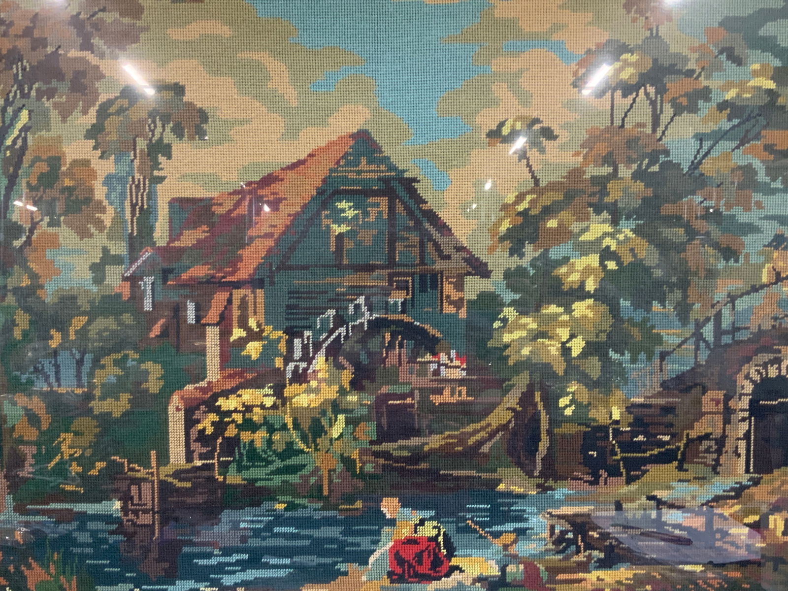 Landscape Cottage Cross Stitch Panel: Vintage cross stitch panel, depicting a landscape with a cottage with a water wheel, a dock with a woman washing clothes in the river Framed in a gold leafed wooden frame with leaf carvings on outer e