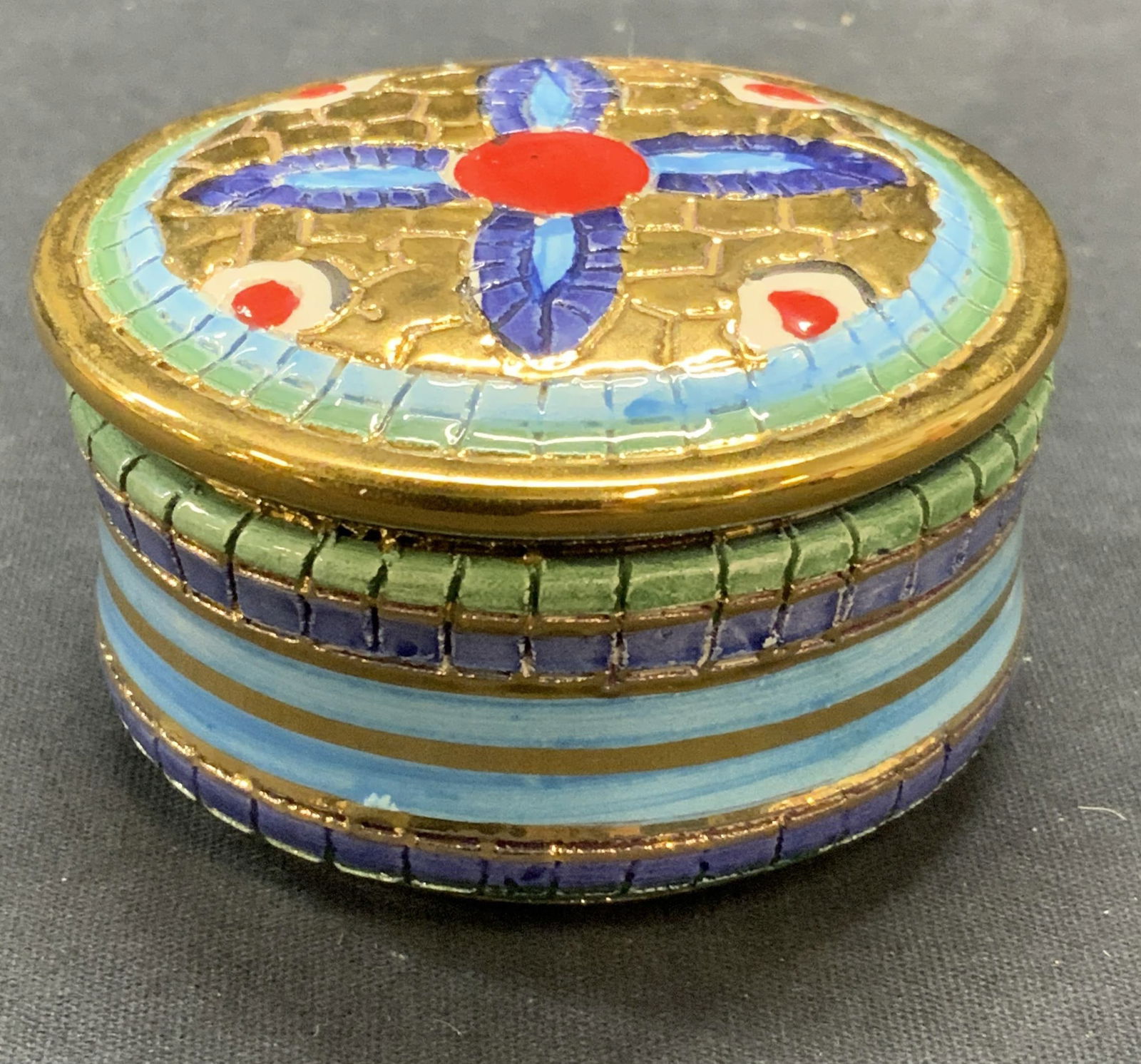 Assisi Gilt Porcelain Trinket Box, Italy: Assisi got porcelain trinket box. Etched surface with mosaic motif. Hand painted in hues of red and blue, signed to underside. Made in Italy. Measures approx. 3 x 1.5 inches. Some surface wear includi