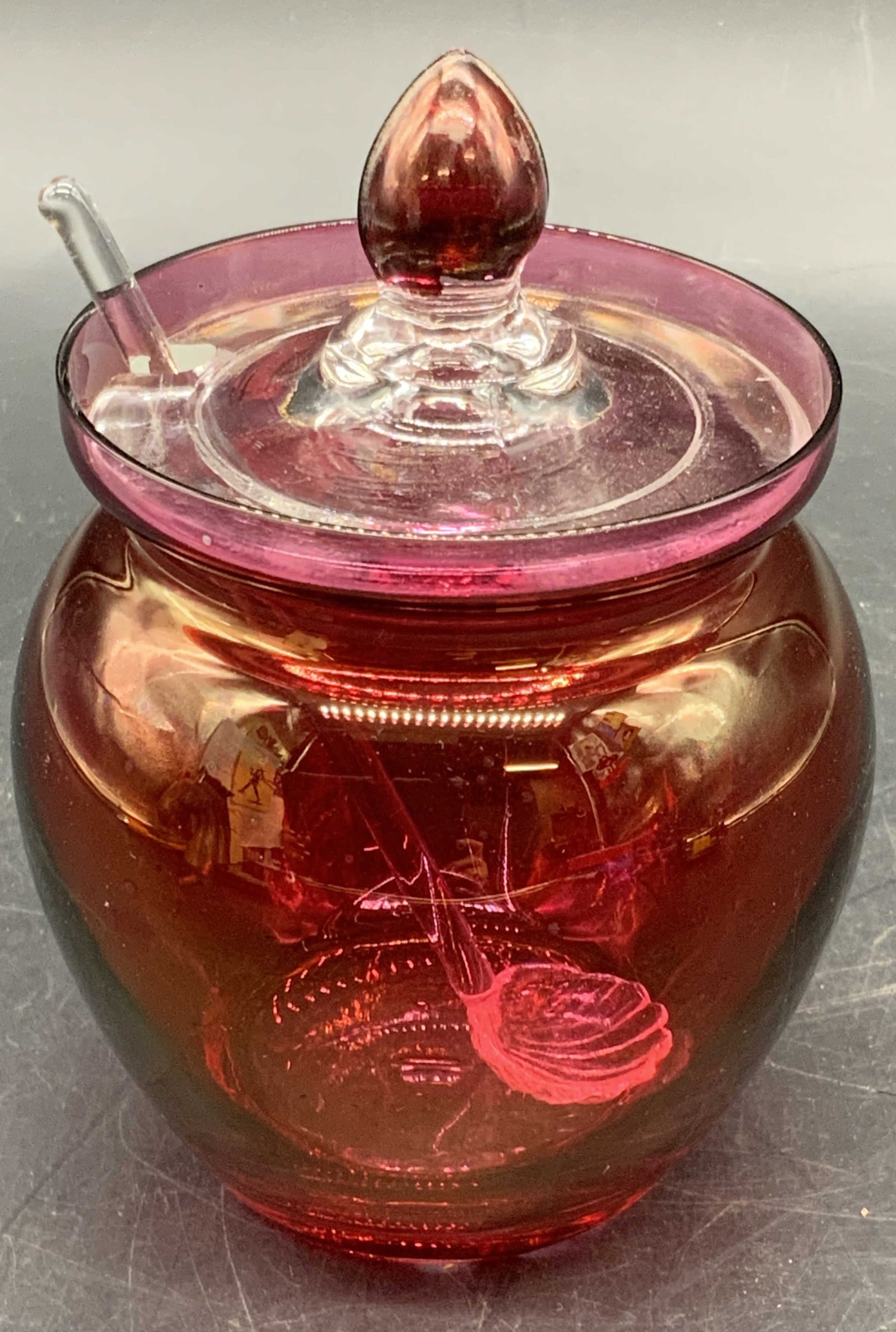 Vintage Cranberry Glass Jam Pot W/ Spoon: A vintage lidded cranberry and clear glass jam pot / jar with a spoon. Finial to the lid. Spoon has a scalloped rim. Jar measures approx. 5H x 3W in. Measured at widest point, measurement includes the