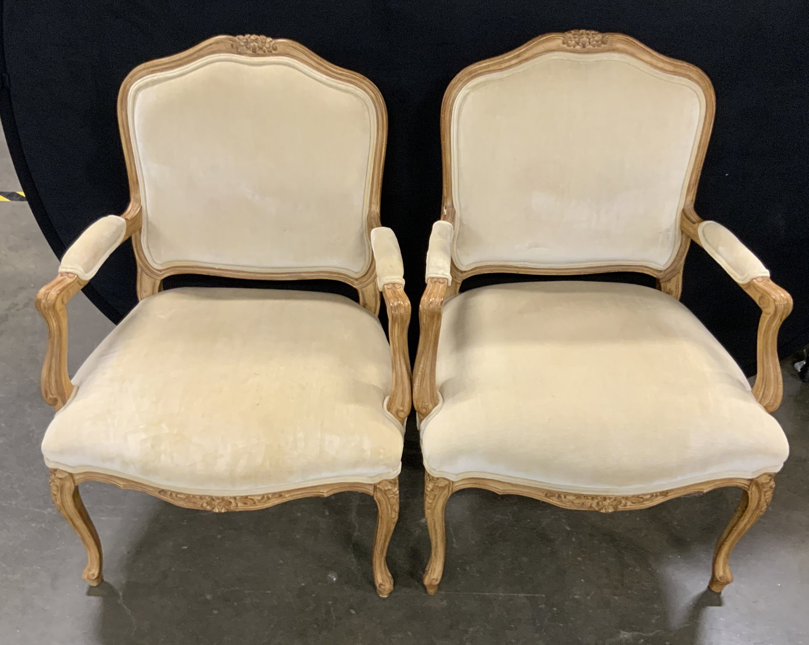 Pair Louis XV Style Vintage Upholstered Armchairs: A pair of vintage Louis XV style upholstered armchairs with carved wooden frames. The chair frames feature floral motifs. Upholstery is soft hand and a light cream hue. Measures approx. 26W x 19D x 38