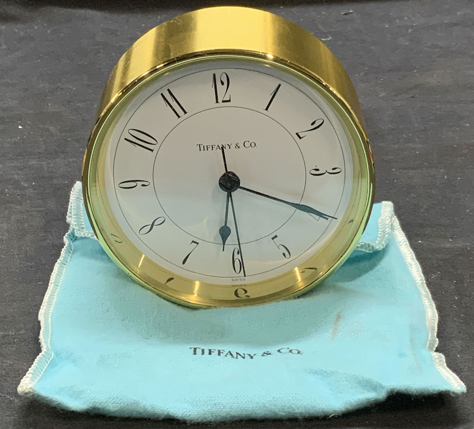 Tiffany & Co Round Brass Swiss Clock: Tiffany & Co round brass Swiss clock. Signed on clock face. Made in Switzerland. Measures approx. 3.75 x 1.5 inches. Includes Tiffany & Co pouch. Piece has weight, not tested for function. Some surfac