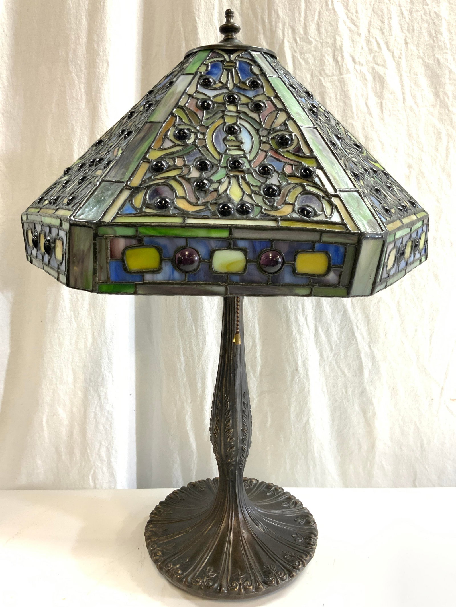 TIFFANY Style Slag Glass Shade Table Lamp: Lamp is Tiffany style. Piece has slag glass lamp shade with multi toned geometric detail. Lamp includes gilt metal finial. Lamp has gilt metal base. Piece has 2 bulb sockets with beaded metal pull swi