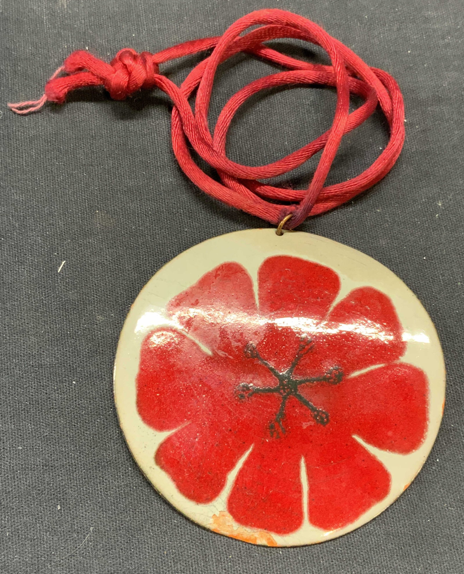 Enameled Copper Flower Pendant Necklace: Pendant necklace with enameled copper pendant and floral motif in red and gray. Red cord. Drop length approx. 12 inches. Pendant measures approx. 3 inches wide. Some surface wear including scratching 