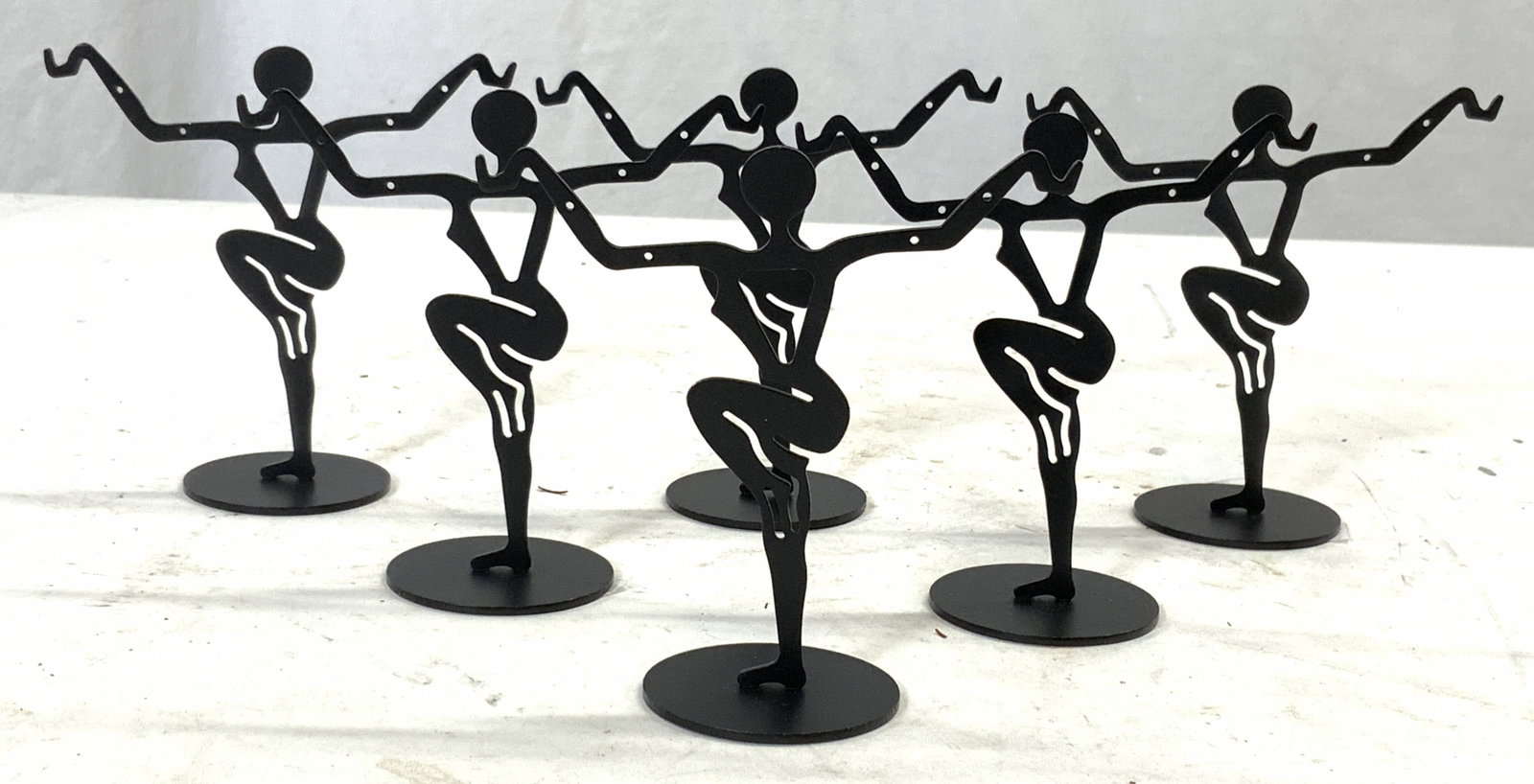Set 6 Metal Female Dancer Figure Earring Holders: Lot includes 6 earring holders. Each are black toned and metal. Each earring holder depicts a female dancer figure with arms up and leg raised. Each piece has circular bases. Each measures approximate