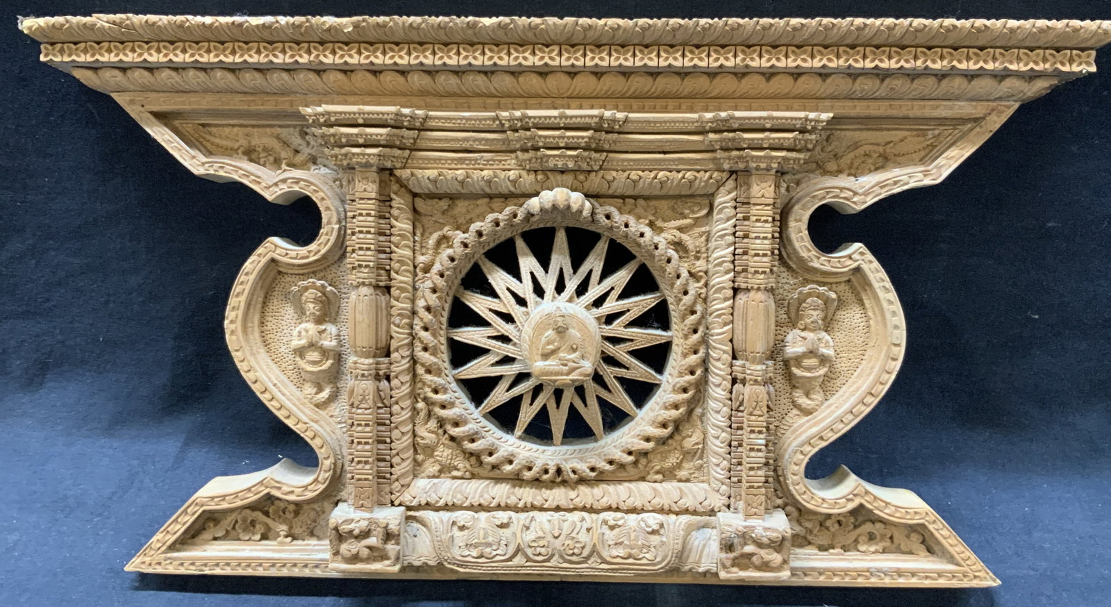 Carved Wooden Buddha Mandala Wall Accessory: Carved tan toned wooden wall paneling depicting a Buddha in the center of a mandala and intricately carved columns and detailing. Approx 8.25 inches tall and 15 inches long Moderate wearing to wood du