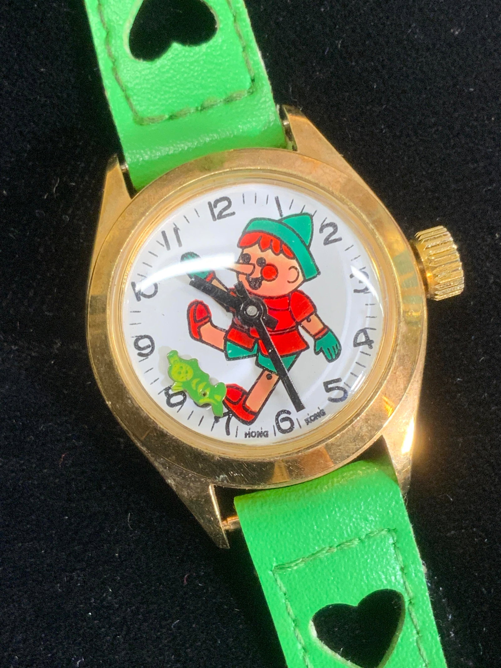 Pinocchio Wristwatch w Green Band: Pinocchio character motif wristwatch with green leatherette wristband that has cut out hearts. Dial watch. 7.5 in. Long. Watch case 1.25 x 1 1/7 in. Loose piece under watch glass. Wristwatch, accessor