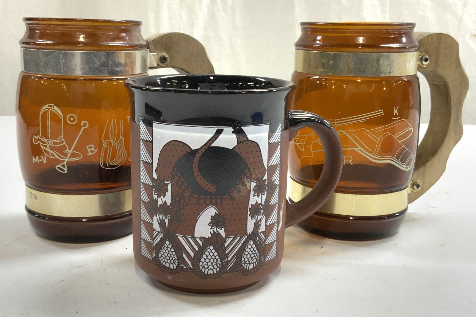 Lot 3 Siesta & Stoneware Mugs Vessels: Underside of each glass mug reads SIESTAWARE. Underside of ceramic mug reads GENUINE ENGLISH STONEWARE. SIESTAWARE mugs are amber glass with cream toned images of firearm, boot, and stirrups, each hav