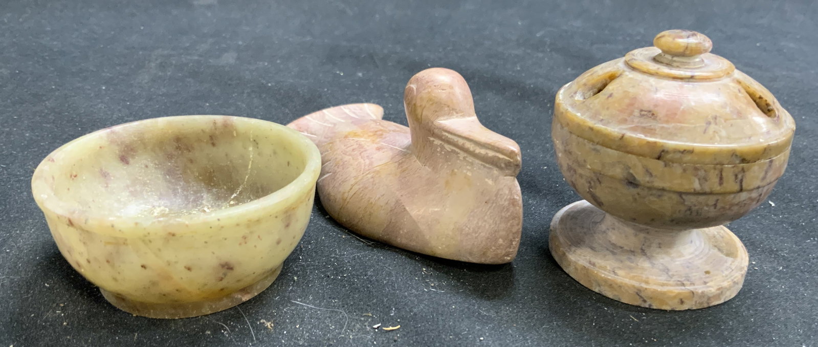 Lot 3 Carved Stoned Vessels & Duck Figure: Lot includes 1 footed vessel with lid, 1 bowl vessel, & 1 carved duck figure. Each piece appears to be carved stone. Duck figure measures approximately 3.75 inches deep 2 inches wide 1.75 inches tall.