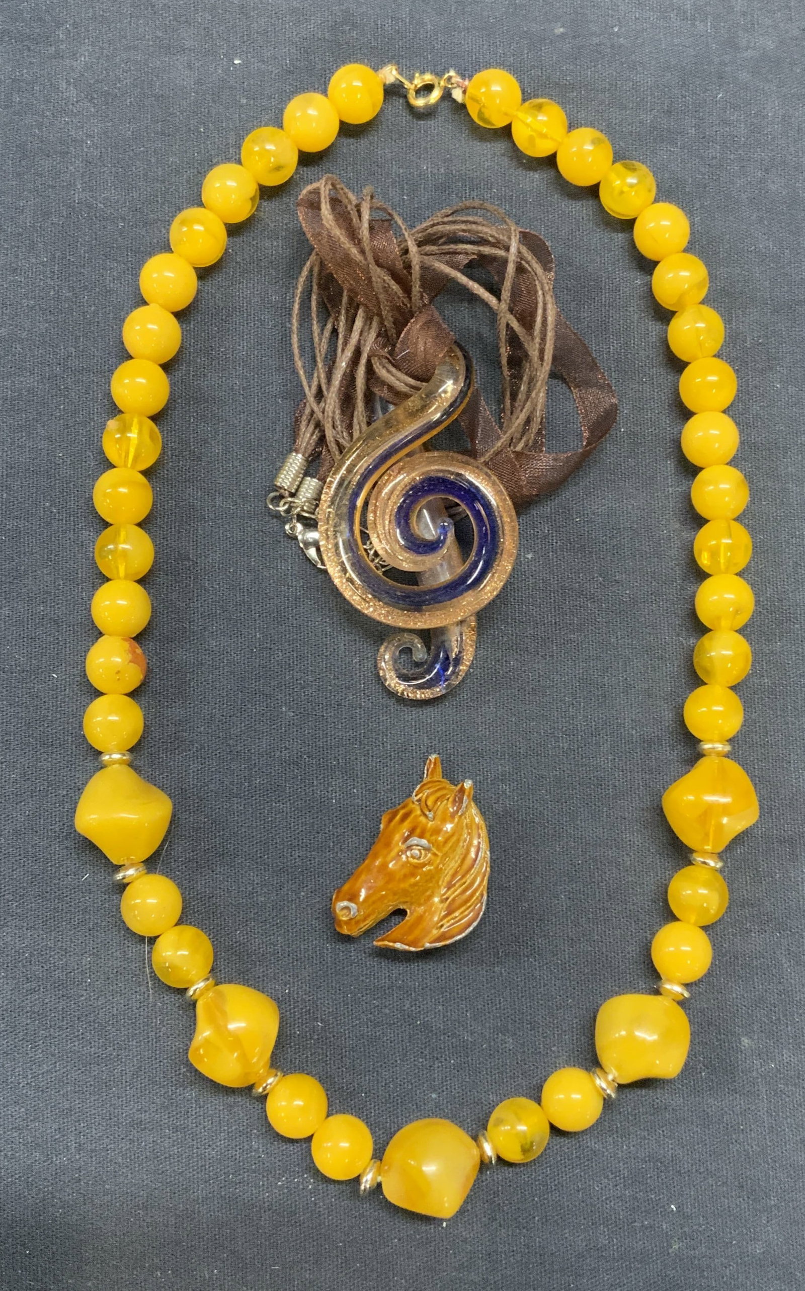 Lot 3 Amber Style Necklace, Horse Brooch +: Amber style beaded lucite necklace. Metallic and blue art glass pendant necklace on brown cord. Enameled Metal horse head brooch. Largest necklace drop length approx. 9.5 inches. Brooch measures appro