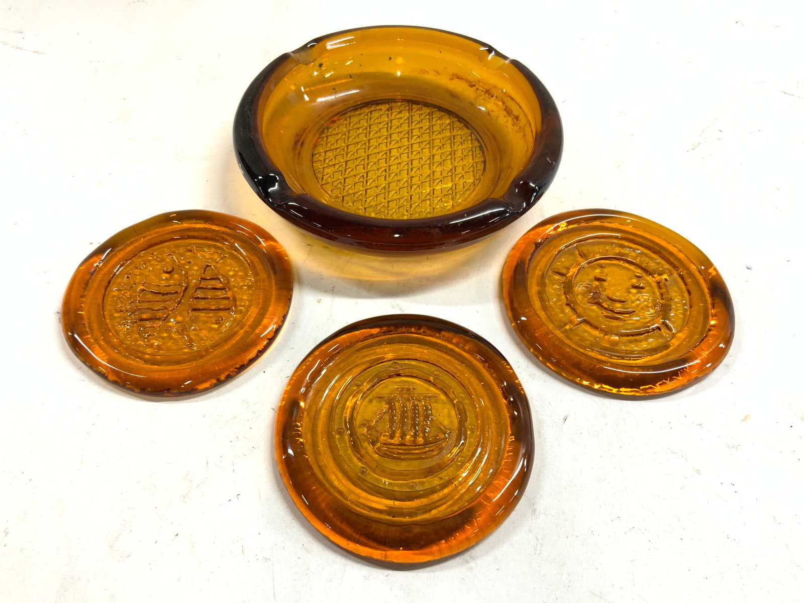 Lot 4 Amber Glass Coasters & Ash Tray: Lot includes 1 ash tray, & 3 coasters. Each piece is amber glass. Coasters have sun, boat, and fish detail. Ash tray measures approximately 6 inches diameter. Ash tray, art glass, amber glass, coaster