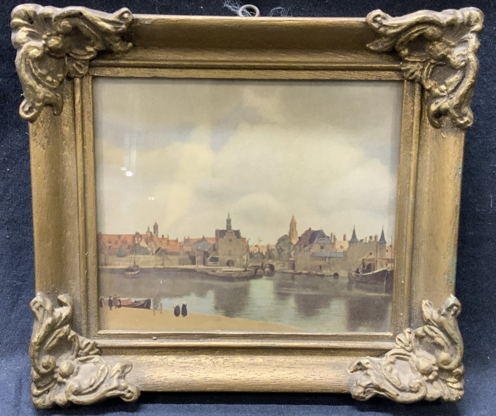 View Of The Delft Lithograph: Lithograph print of the view of the Delft, depicting the view of the waterway with the city in the distance. Framed in gold leafed wooden frame with ornate corners. Approx 6 x 7 inches window measures