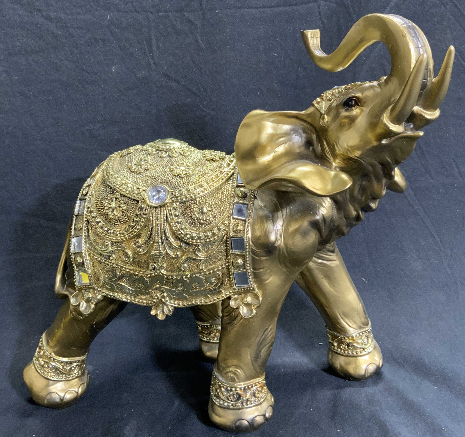 Resinous Gold Tone Elephant Figure: Elephant figure appears to be crafted from a resinous material. Piece is gold toned. Depicts a standing elephant with detailed saddle & trunk raised. Piece has some mirrored surfaces. Measures approx 